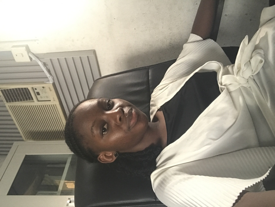 The image captures a young Black female seated in a black office chair, looking directly at the camera with a neutral expression. She wears a black top under an off-white, pleated, short-sleeved wrap garment tied at the waist, and dark stud earrings. Her hair is dark and styled in braids. The background depicts an interior room with a light-toned wall. To the subject's left (viewer's right), a white-framed window or glass panel is partially visible. Above and to the subject's right (viewer's left), an LG brand window air conditioning unit with horizontal vents and a control panel is mounted on the wall. A white electrical switch or outlet, connected by a diagonal white conduit, is positioned on the wall to the right of the air conditioner. The wall exhibits minor discoloration. No specific visual cues are present within the image to determine the precise location in the city of Ado, Nigeria.