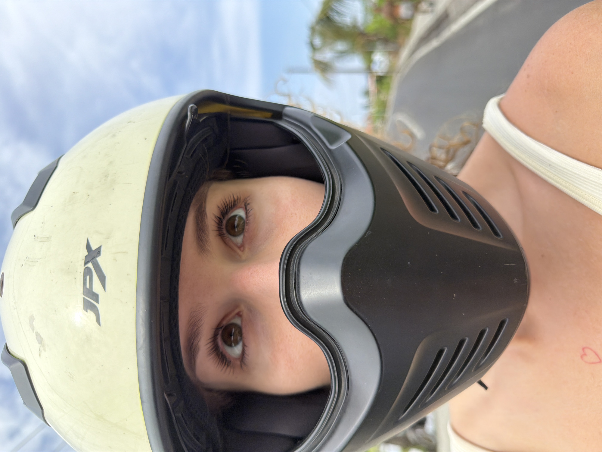 A close-up, forward-facing, medium shot captures the upper body and face of a person wearing a light yellow helmet with black trim, and a black face shield. The person's visible face consists of their eyes, eyebrows, and a portion of their cheeks. The eyes are brown and wide open, looking upwards and slightly to the left. Long, dark eyelashes are prominent. The helmet has a grey logo that reads "JPX". The face shield is black and covers the lower half of the face, featuring a vented design. The person is wearing a cream-colored tank top. A faint red heart tattoo is visible on the left collarbone. The background is blurred and depicts a road with palm trees and foliage, suggesting an outdoor, possibly tropical, environment. The sky is blue with scattered white clouds. The scene appears to be captured while moving, as indicated by the motion blur of the background. The location is identified as the city of Mengwi, Indonesia.