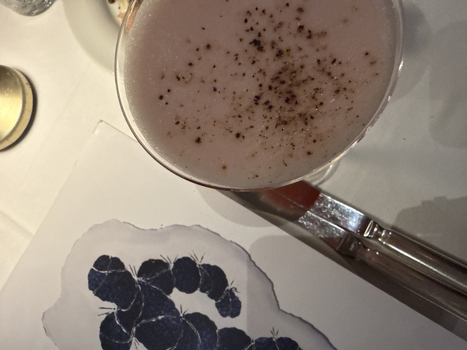 A cocktail with a light pink frothy surface and dark specks rests near a place setting. Adjacent to the glass is a silver fork or pair of tongs. Below, a white card with a blue cactus design lies partially visible. A round golden lid is present on the upper left side of the frame. The backdrop appears to be a white tablecloth. The scene suggests a restaurant or bar setting.