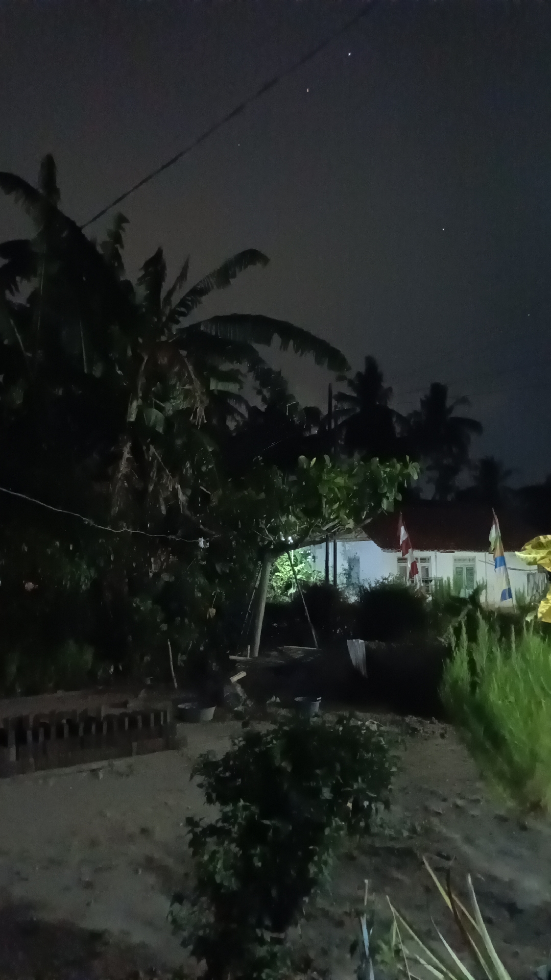 A nighttime, outdoor, low-light scene in Kebumen, Indonesia, depicts a dimly lit yard with several trees and a house in the background. Banana trees are prominent on the left side of the frame. A building with a red roof and white walls, possibly a house, is visible in the mid-ground. Two flags are attached to poles near the house. The ground is composed of dirt and sand. In the sky, several small, bright points of light are visible, which could be stars or distant lights. The overall atmosphere is dark and rural.