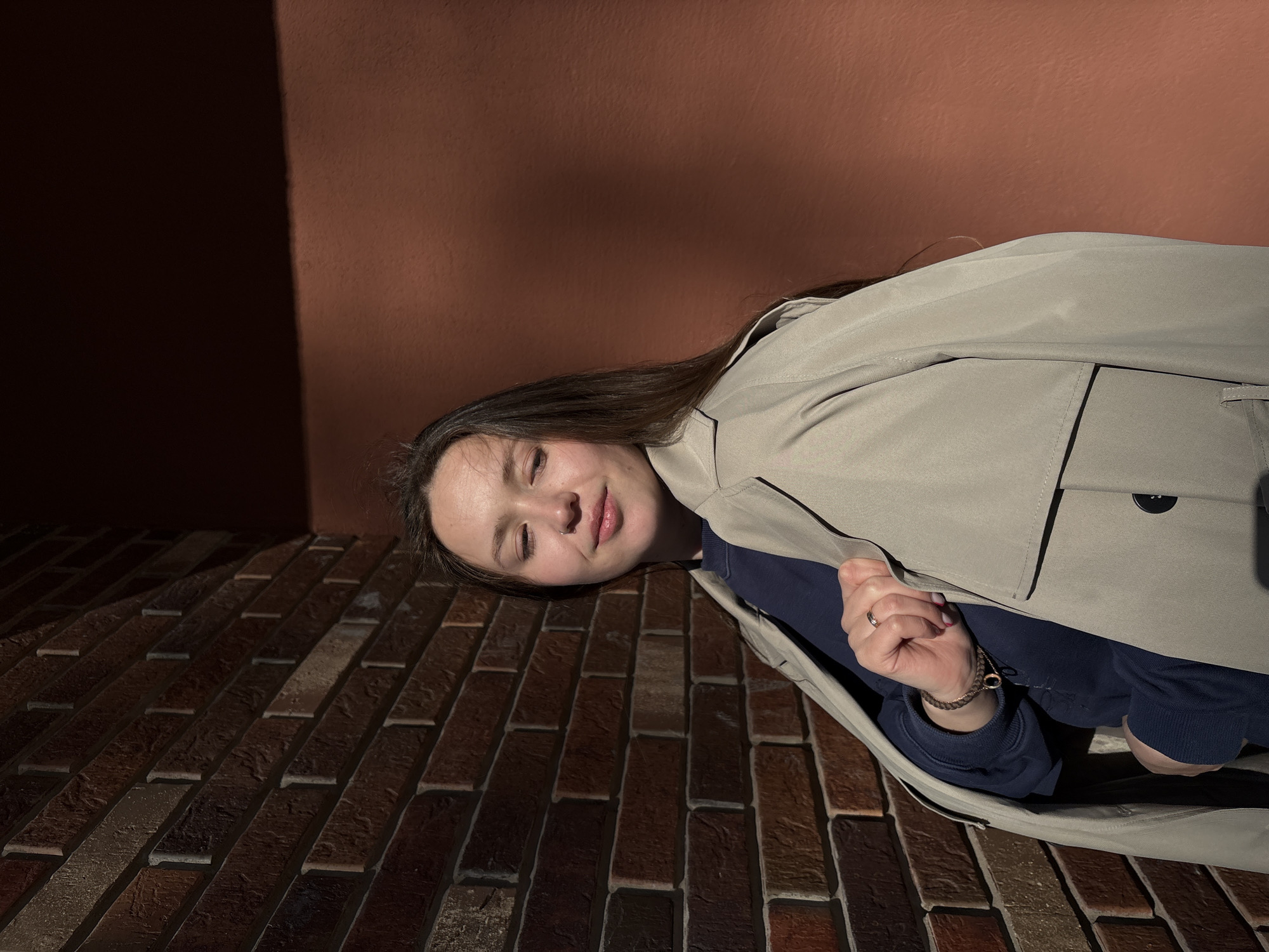 A young woman in a trench coat lies on a brick surface next to a wall. The wall is split into dark and light brown sections, with a sharp shadow line dividing them. The brick surface is made of dark reddish-brown bricks arranged in rows. The woman is partially covered by the trench coat, but her face, hand, and part of her arm are visible. She wears a dark blue long-sleeved shirt. A ring is visible on her hand, and a bracelet is around her wrist. The scene is illuminated by direct sunlight. The location is Izhevsk, Russia.