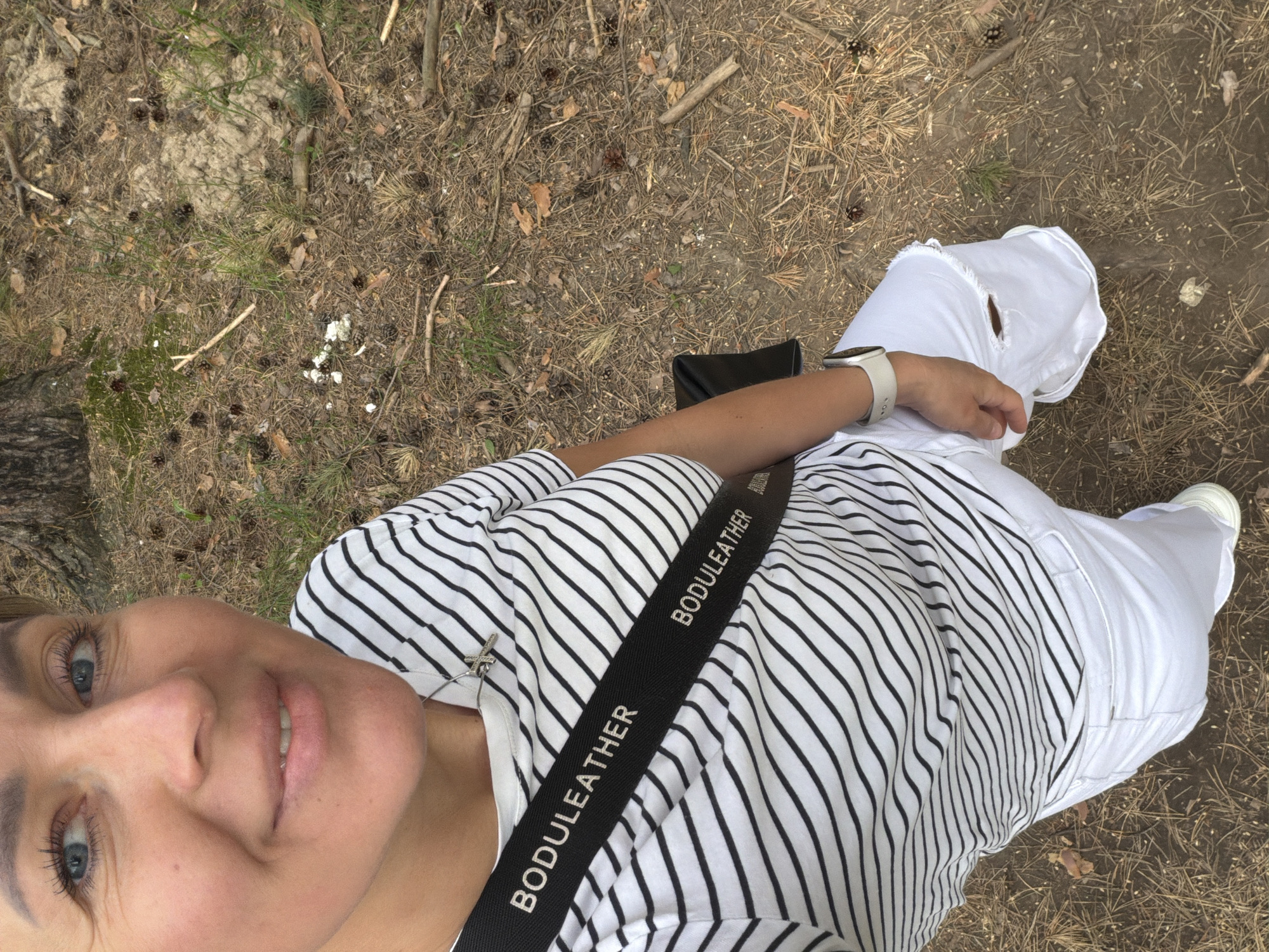A selfie shows a woman in a park setting. She wears a striped top, white pants with a hole in the knee, and white sneakers. She has a black crossbody bag with the inscription "BODULEATHER" on the strap and a light-colored watch on her wrist. The ground is covered with dry leaves and pine needles, with some patches of grass and a tree trunk visible on the left. The location is Chaikovsky, Russia.