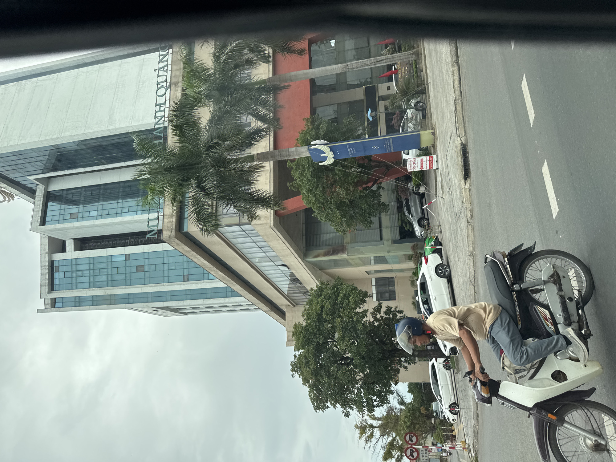 A man rides a motorcycle on a street in Nam Đông Hà Ward, Vietnam. The man, wearing a light brown t-shirt and blue pants, has a blue helmet and is looking forward. His motorcycle is dark brown and white. In the background, a tall, modern building with blue-tinted glass windows stands against a cloudy sky. Palm trees and other green foliage are visible in front of the building. Several cars, including white sedans and a silver SUV, are parked or driving on the street. Traffic signs and a blue directional sign are also present.