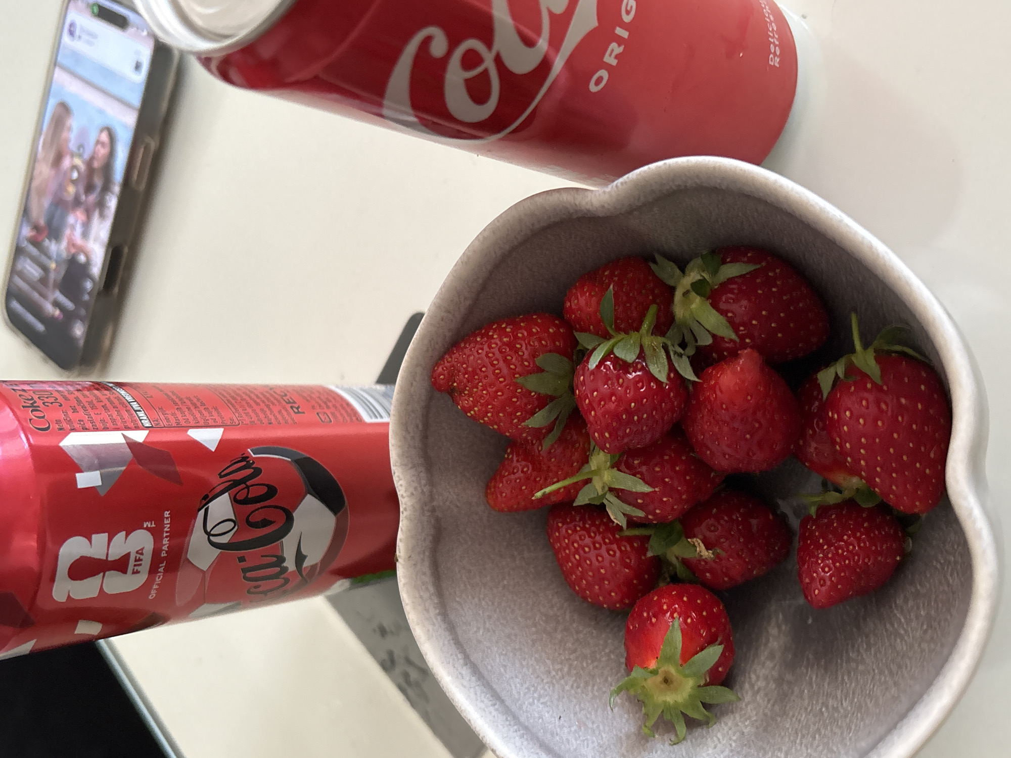 The scene features a table with a bowl of strawberries, two cans of Coca-Cola, and a smartphone. The strawberries are bright red and have green stems, filling a grey heart-shaped bowl. One can of Coca-Cola is upright, displaying the classic logo, while the other can is horizontal and shows FIFA World Cup promotional branding. A smartphone lies on the table displaying what appears to be a photograph of three women. The setting is likely indoors, given the neutral-colored table surface and soft lighting. There is no explicit indication of Kragujevac, Serbia.
