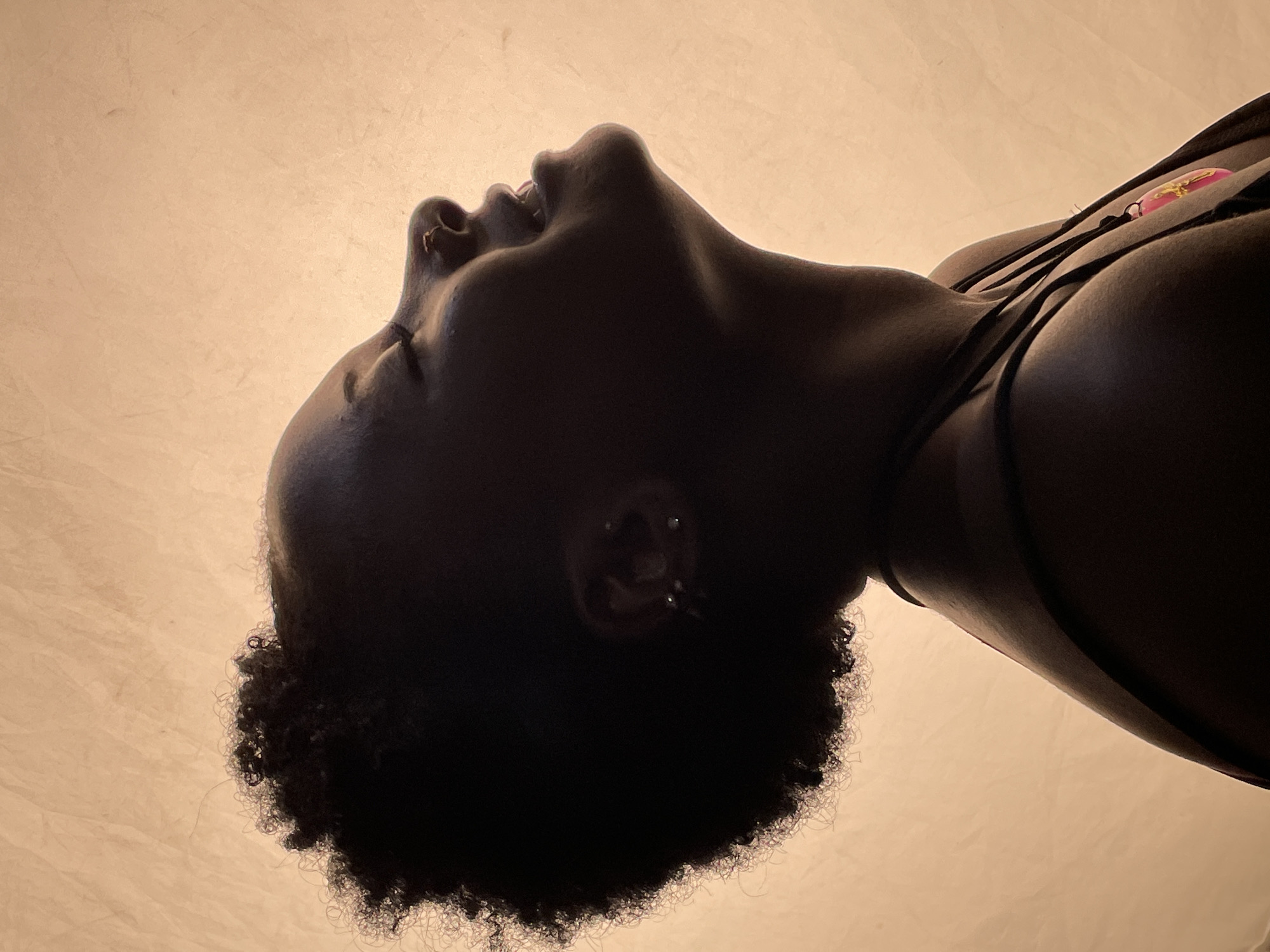 A low-angle shot depicts a dark-skinned individual with tightly curled hair, head tilted back, facing upwards. The person appears to be wearing a dark top or tank top. The background is a neutral, light-colored surface, possibly a ceiling or wall. The setting is unknown and cannot be determined to be in Jos, Nigeria.