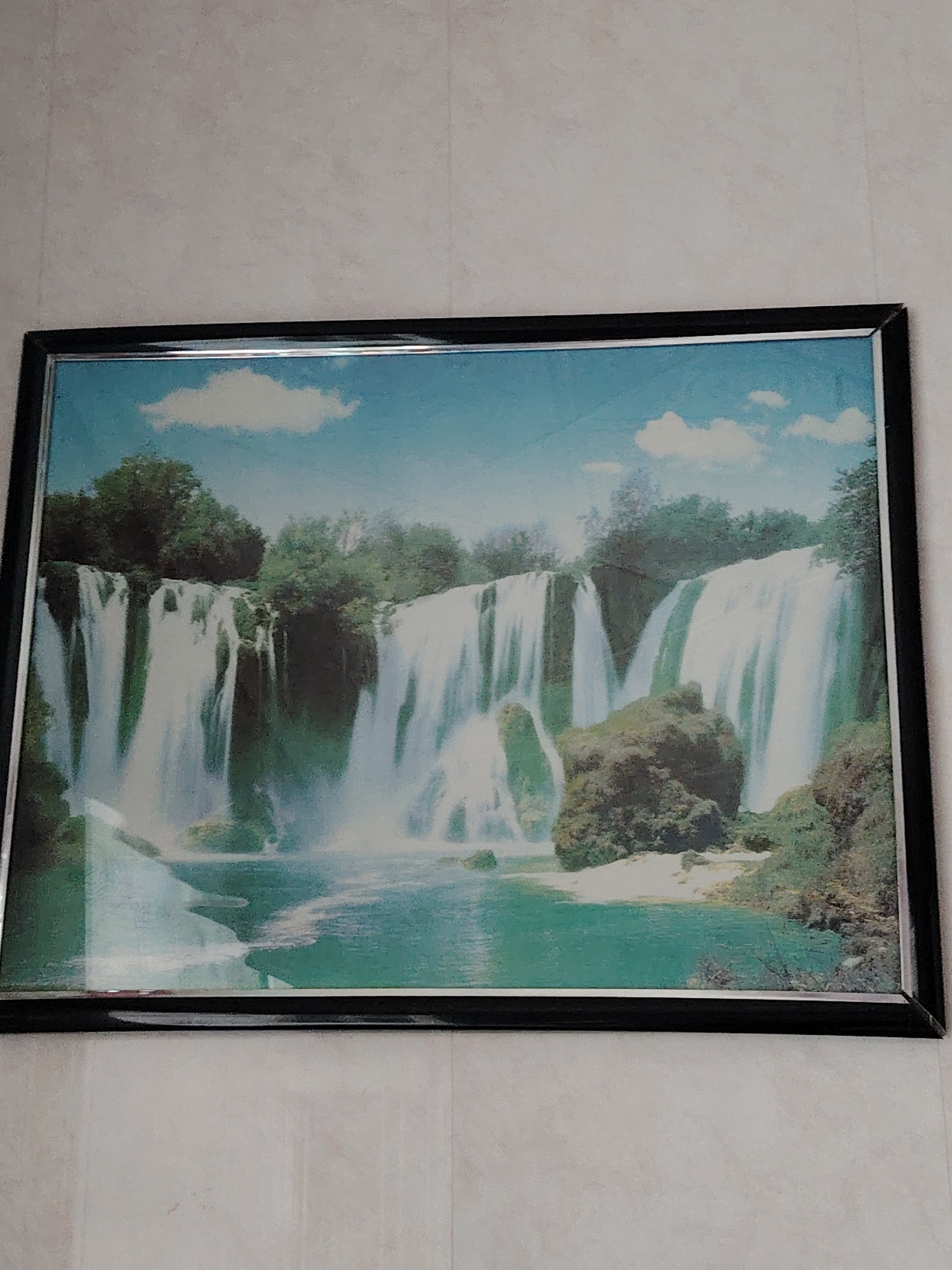 The image displays a framed picture of a large, multi-tiered waterfall, hung on a textured, light-colored wall in Yanaul, Russia. The picture, oriented horizontally, is encased in a simple black frame with a thin silver inner border.
Within the frame, a vibrant natural landscape is depicted. Numerous cascades of white water plunge from a green, tree-covered cliff into a wide, clear turquoise body of water at the base. Lush green foliage and trees densely cover the top of the cliffs and the banks surrounding the water. Large rocks and boulders are visible within the waterfall's path and along the water's edge. The sky above is a bright blue, dotted with several soft, white cumulus clouds. The lighting suggests a bright, sunny daytime scene.
The wall on which the picture hangs appears to be light beige or off-white, with a subtle vertical striation pattern. There are no people visible in the image, nor any specific activities taking place other than the static display of the artwork. No text is visible on the picture or its frame. The overall impression is one of serene natural beauty, captured and presented indoors.