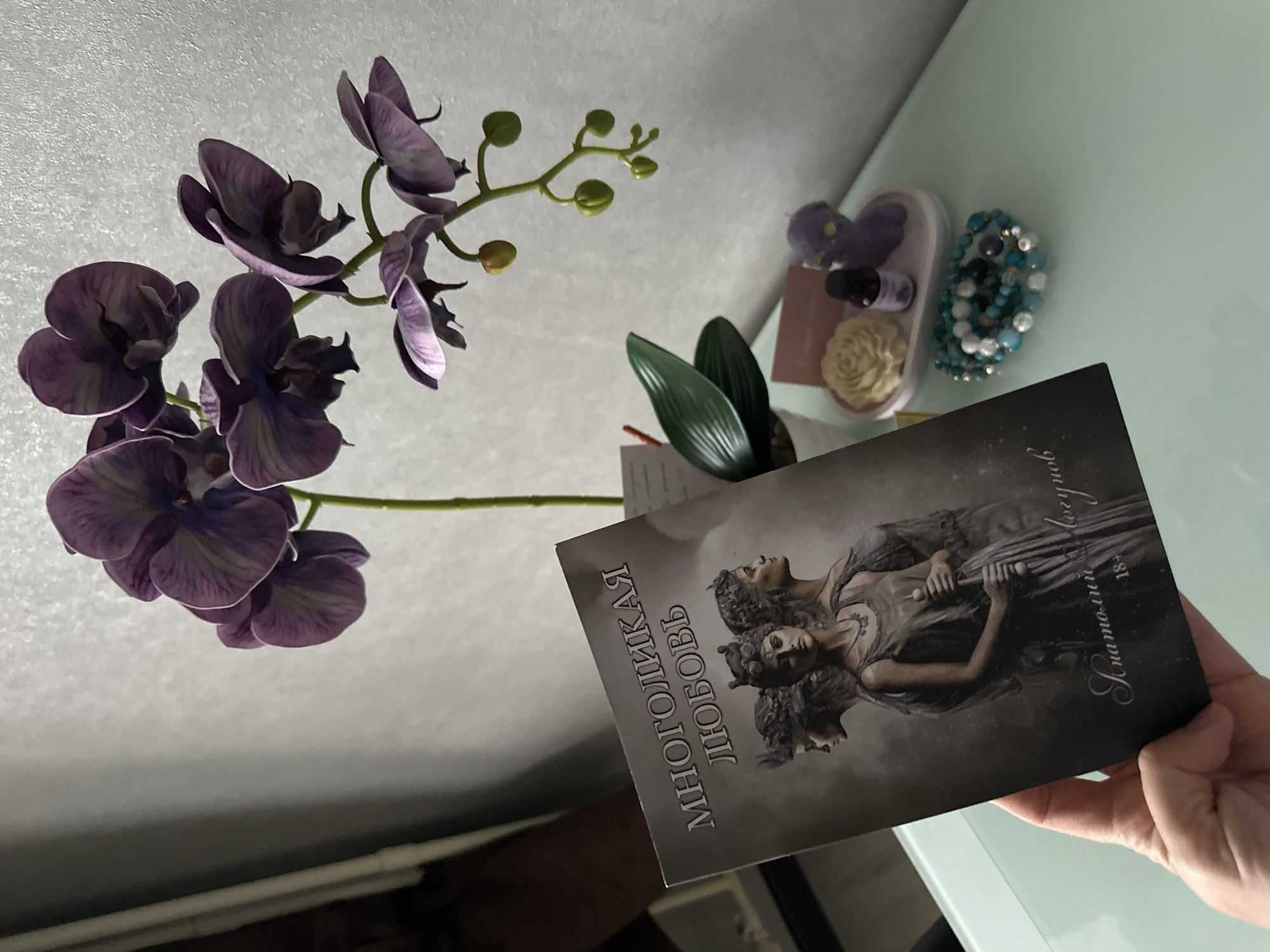 A person's hand holds a book with a dark cover, featuring the title "МНОГОЛИКАЯ ЛЮБОВЬ" (Many-Faced Love) in Cyrillic script. The book's cover depicts three women with elaborate hairstyles, rendered in a grey, atmospheric style. Below the title, the author's name, "Анатолий Вогулов," is written in a cursive font, followed by "18+". To the right of the book, a potted artificial orchid with purple flowers and green leaves stands on a light blue surface. Beside the orchid, on a small tray, are a yellow rose-shaped object, a small dark bottle, and a purple figurine. A stack of beaded bracelets in shades of blue, white, and silver rests on the surface in front of the tray. The background is a plain, light-colored wall with a textured finish. The scene appears to be indoors, likely on a table or desk.