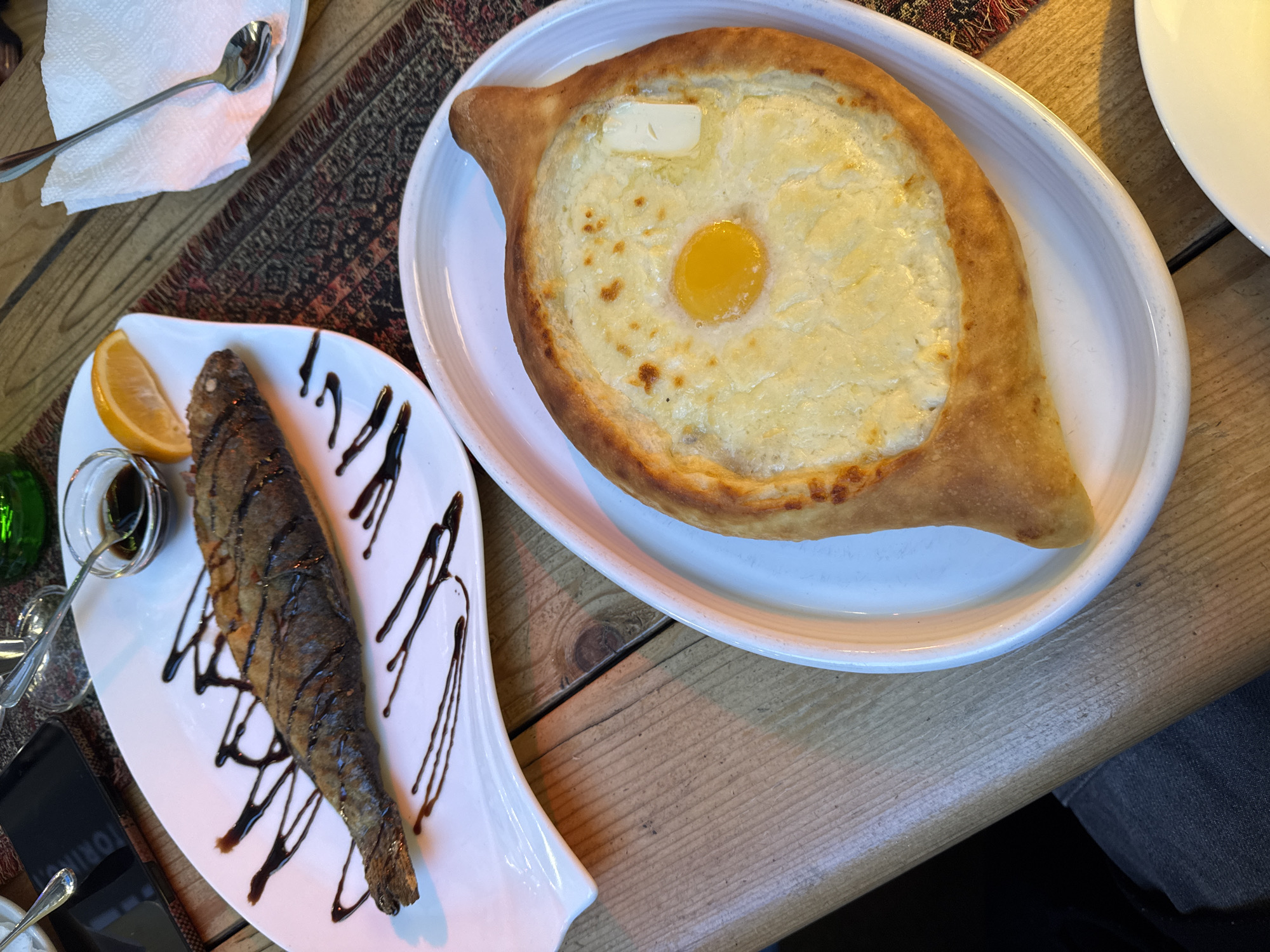 The image shows two dishes presented on a wooden table, presumably in a restaurant in Tbilisi, Georgia. On the left is a plate with a fried fish, drizzled with a dark sauce, accompanied by a lemon wedge. A small glass container with a dark liquid and a spoon is placed near the fish. On the right, another plate holds an Adjaruli Khachapuri, a Georgian bread dish filled with cheese and topped with an egg yolk and a pat of butter. A spoon and a patterned placemat are visible in the upper left corner of the image. A portion of a white plate is seen to the right of the Khachapuri. A section of a person's leg wearing denim is visible on the lower right of the image.