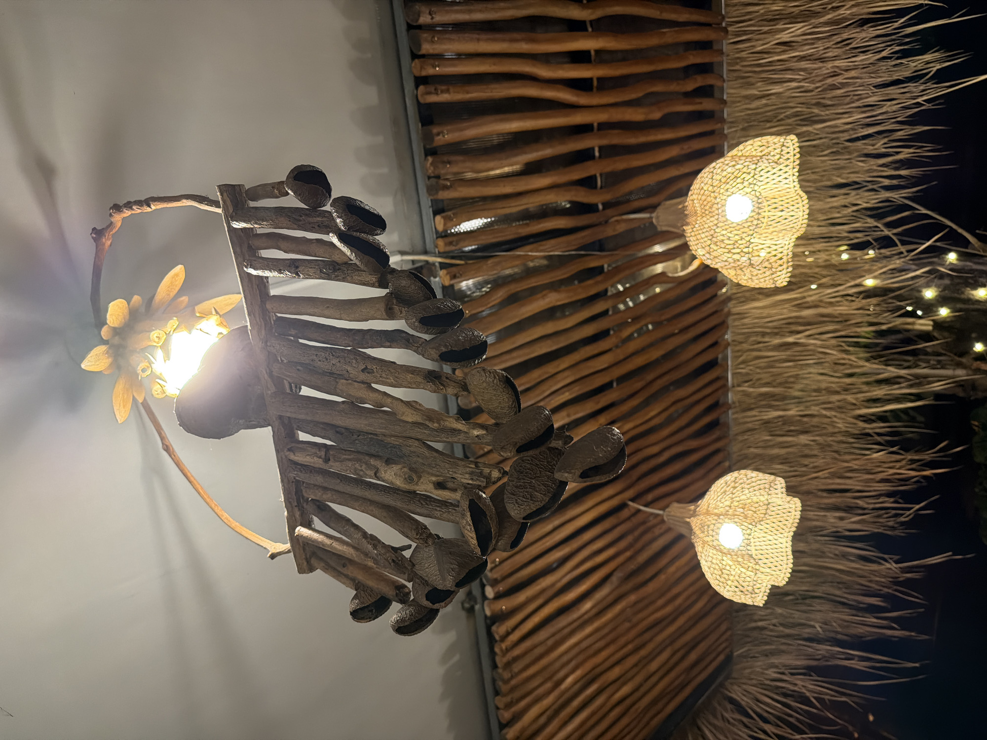 Captured in North Kuta, Indonesia, the image features two distinct lighting fixtures affixed to a ceiling. On the left, a chandelier-like structure is constructed from dark wood, featuring hollow seed pods hanging from a frame of branches. The central light source emits a warm glow, illuminating the chandelier's wooden components. To the right, two more lights are visible, enclosed in woven shades resembling stylized flowers. These lights emit a softer, diffuse light. The ceiling itself has varied textures, including a section made of horizontally arranged twigs and a thatched area with long, straw-like material. Tiny, twinkling lights are subtly visible in the background, behind the thatched section of the ceiling.