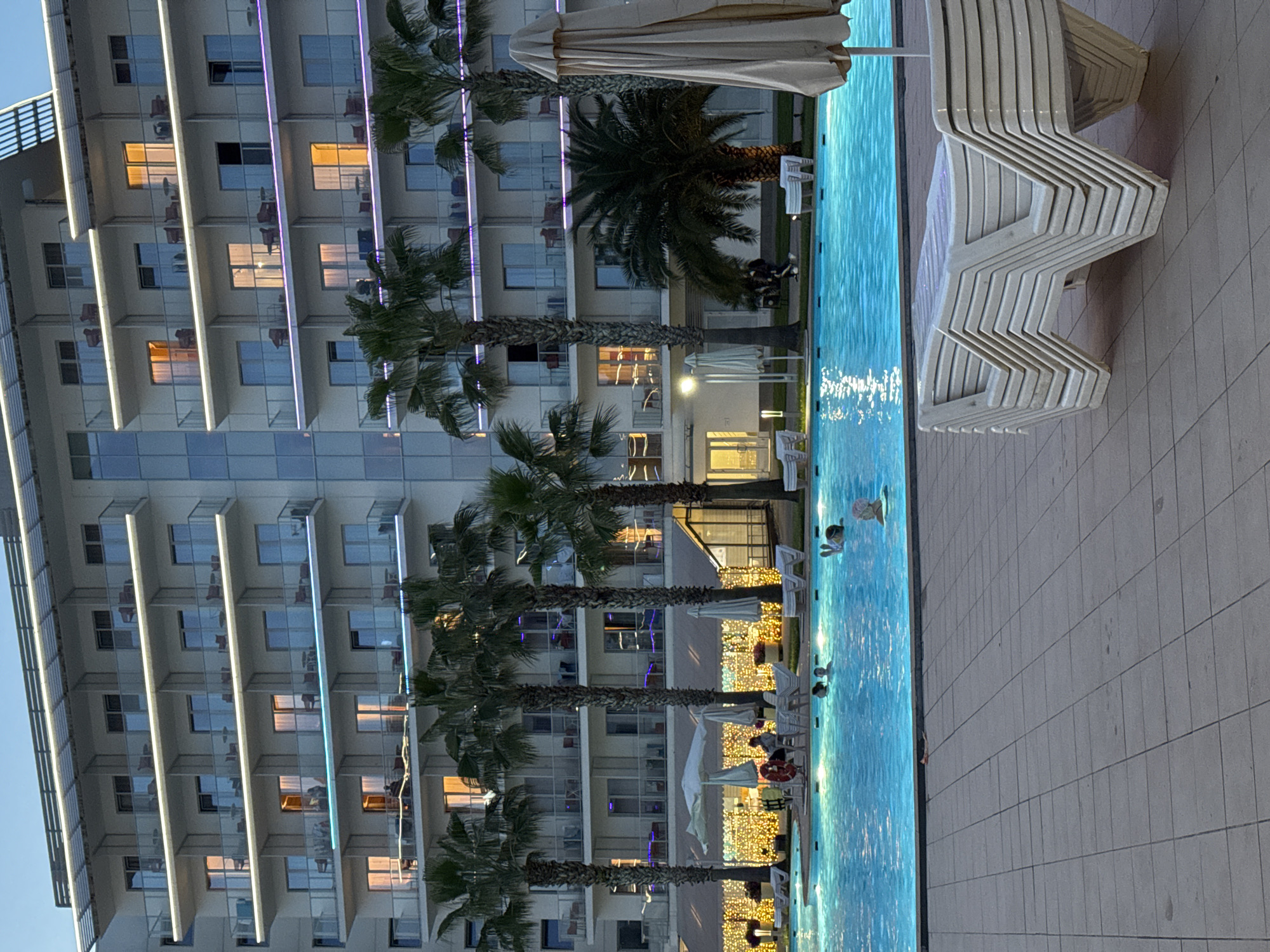 The image captures a multi-story hotel in Sochi, Russia, situated beside a swimming pool. The hotel features numerous balconies and illuminated accent lights on each floor. Several palm trees line the perimeter of the pool. In the pool, there are multiple people swimming. Beside the pool are stacked lounge chairs, and further down, sun umbrellas are seen. The area around the pool is tiled.