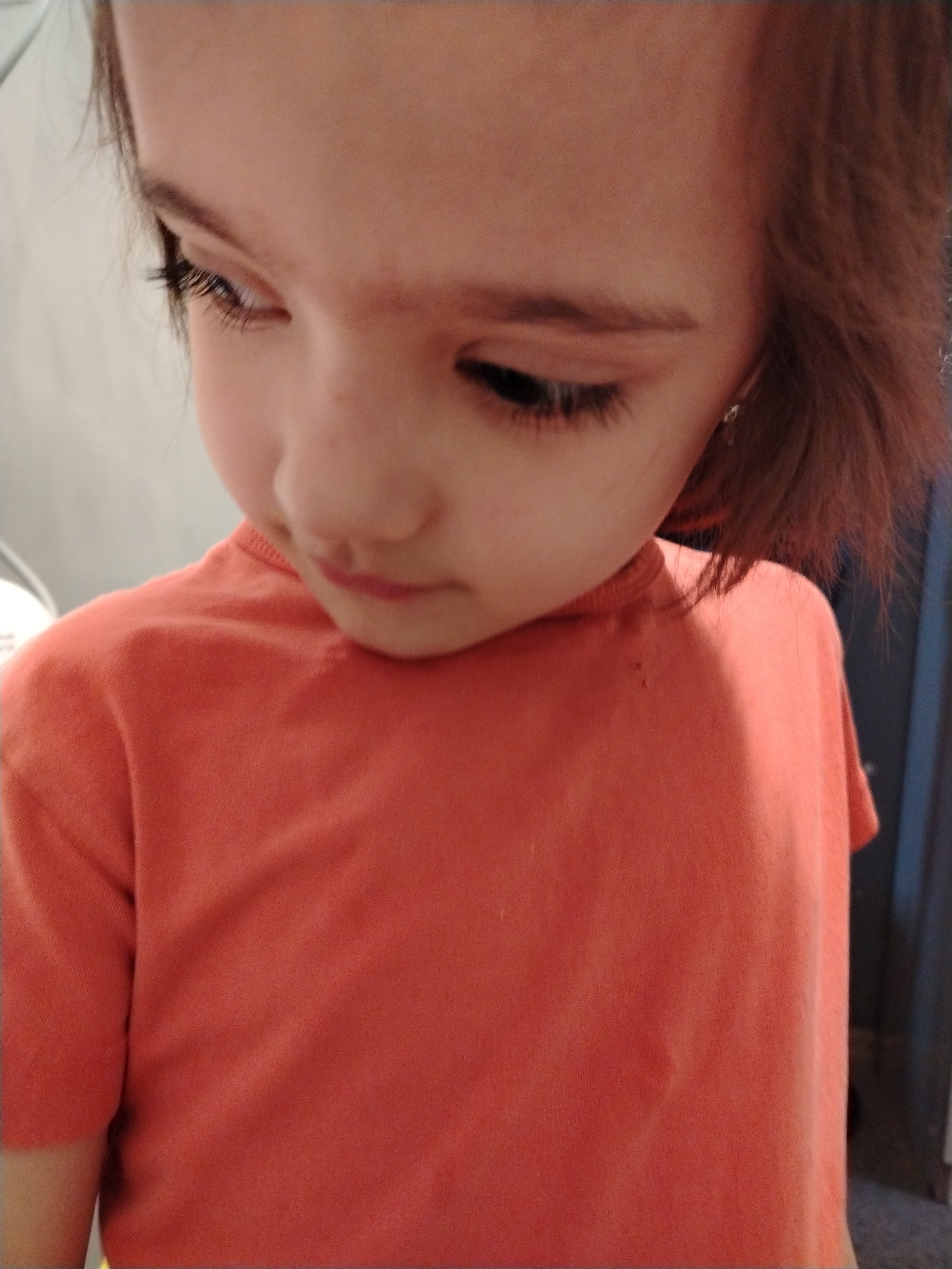 A young child with brown hair and an orange short-sleeved shirt is looking down. The background is blurry and shows parts of a wall and what appears to be a door. The child's face is the primary focus, capturing a moment of introspection. The location of the photo within the city of Noviy, Russia cannot be determined from the image alone.