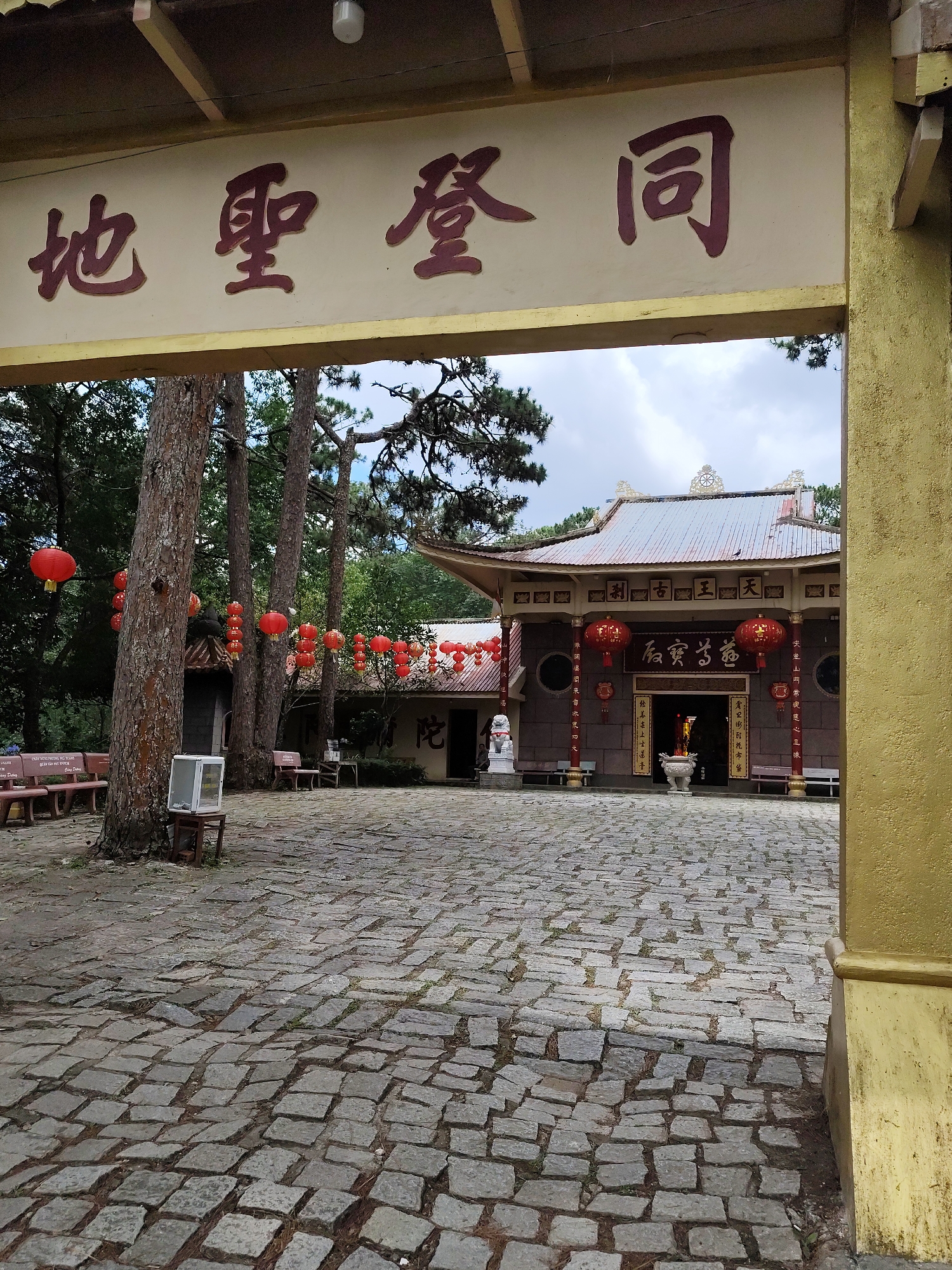 Captured through a weathered gate in Da Lat, Vietnam, the image showcases the courtyard of a Buddhist temple. The courtyard is paved with stone blocks in a haphazard pattern. Red paper lanterns are strung between trees and along the roofline of the building. The temple features a metallic roof, stone walls, and circular windows, along with signage in both Chinese and Vietnamese. A lion statue stands guard near the entrance. Two sets of benches provide seating. A small, boxy appliance sits on a stool near a tall tree. The sky is overcast.