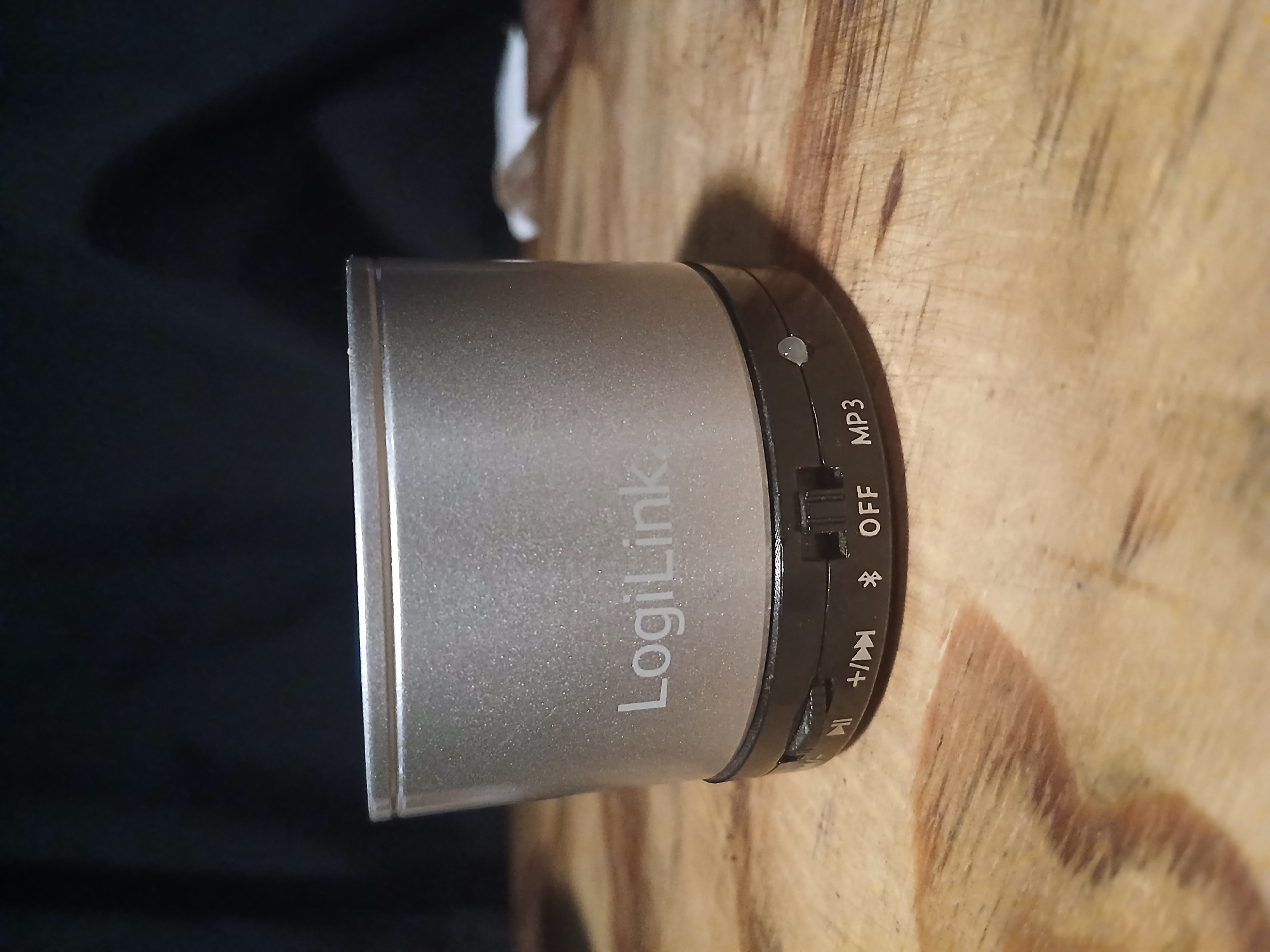 A silver cylindrical speaker with "LogiLink" printed on it sits on a wooden surface. The speaker has a black base with several control buttons: a play/pause button, a volume up/next track button, a Bluetooth icon, an "OFF" setting, and "MP3". The background is dark and out of focus. The image does not contain any information about the city of Douai, France.