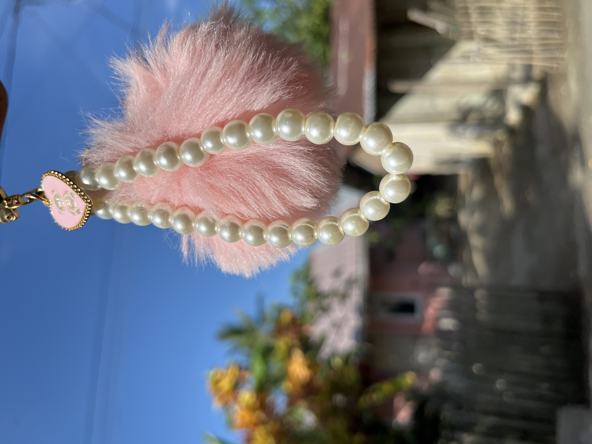 A close-up shot focuses on a pink fluffy pom-pom adorned with a string of pearls and a heart-shaped charm featuring a teddy bear. The pom-pom and pearls are suspended in the foreground, appearing to hang from a window or similar vantage point. In the blurred background, a rural Indonesian scene is visible, with a wooden fence, trees with orange blossoms, and simple houses under a clear blue sky. The overall impression is one of a decorative item set against a backdrop of everyday life in a tropical location.