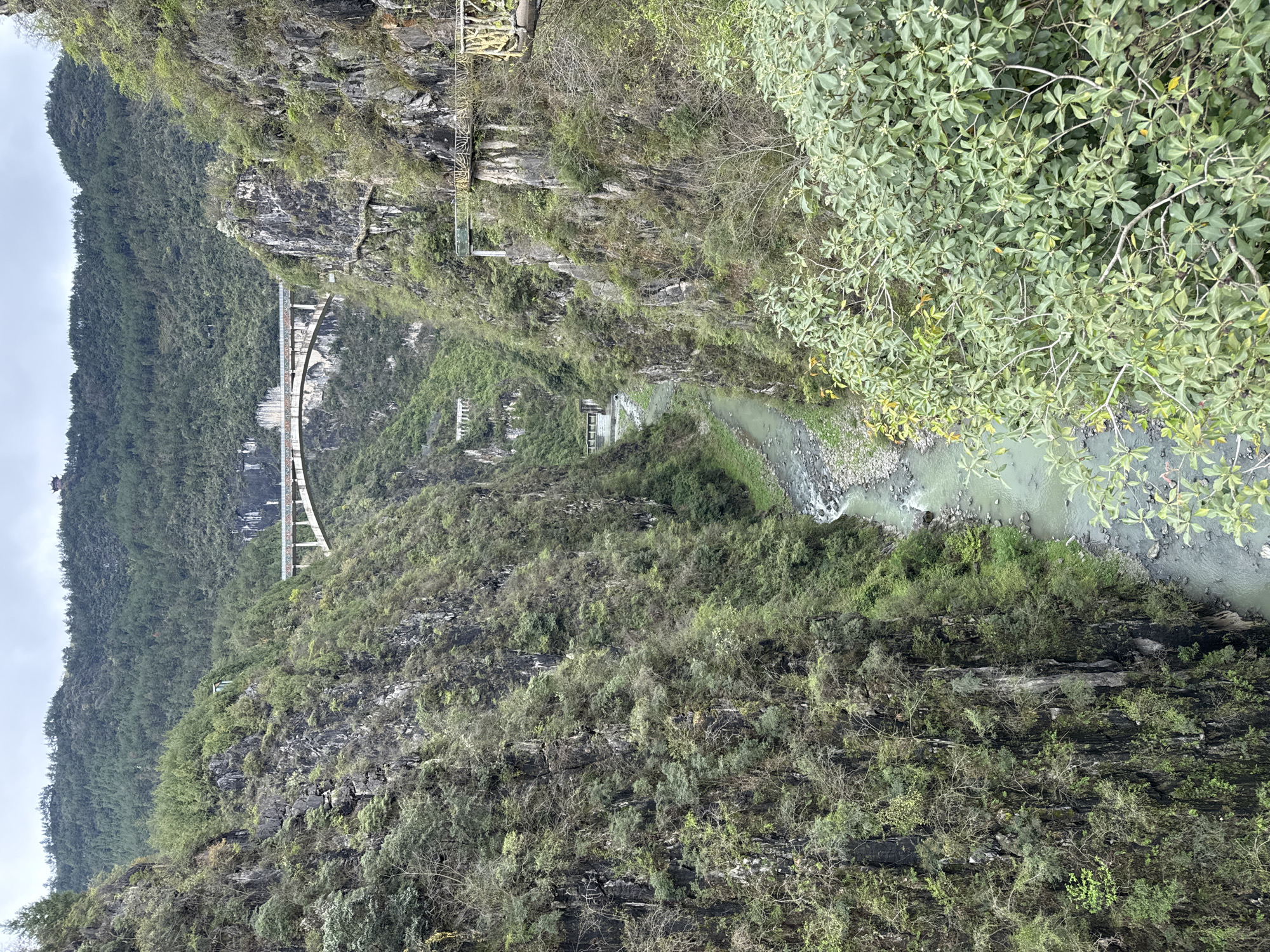 A view of the Aizhai Bridge in Qianjiang, China. A river flows through a ravine between two mountain sides. The bridge is in the distance. The mountains are covered in dense vegetation. The sky is overcast. A catwalk structure is located along a cliff.