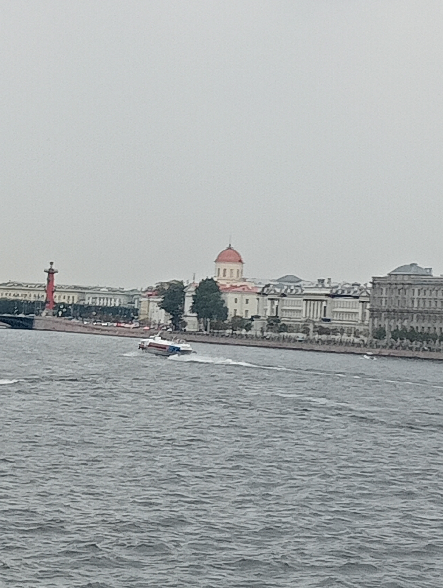 This is a medium shot taken from a boat or from a low vantage point, looking across a body of water toward a city skyline. The sky is overcast and a uniform gray.
In the foreground, the water is a choppy, gray-blue, with whitecaps and ripples suggesting movement. A white and red speed boat with blue stripes is visible in the middle of the frame, moving from left to right and leaving a wake. The boat appears to be a passenger ferry or similar vessel.
The background shows a waterfront lined with a series of buildings. On the far left, a tall, red, columnar structure, possibly a lighthouse or monument, stands prominently. To its right are several large, classical-style buildings with ornate facades and columns. One building has a distinctive red dome. Trees are visible along the waterfront, and a pedestrian area or walkway appears to run in front of the buildings. The overall architectural style suggests a historic European city.
This scene is likely the Spit of Vasilievsky Island in Saint Petersburg, Russia, a famous landmark featuring the Rostral Columns and the Old Stock Exchange Building, viewed from the Neva River.