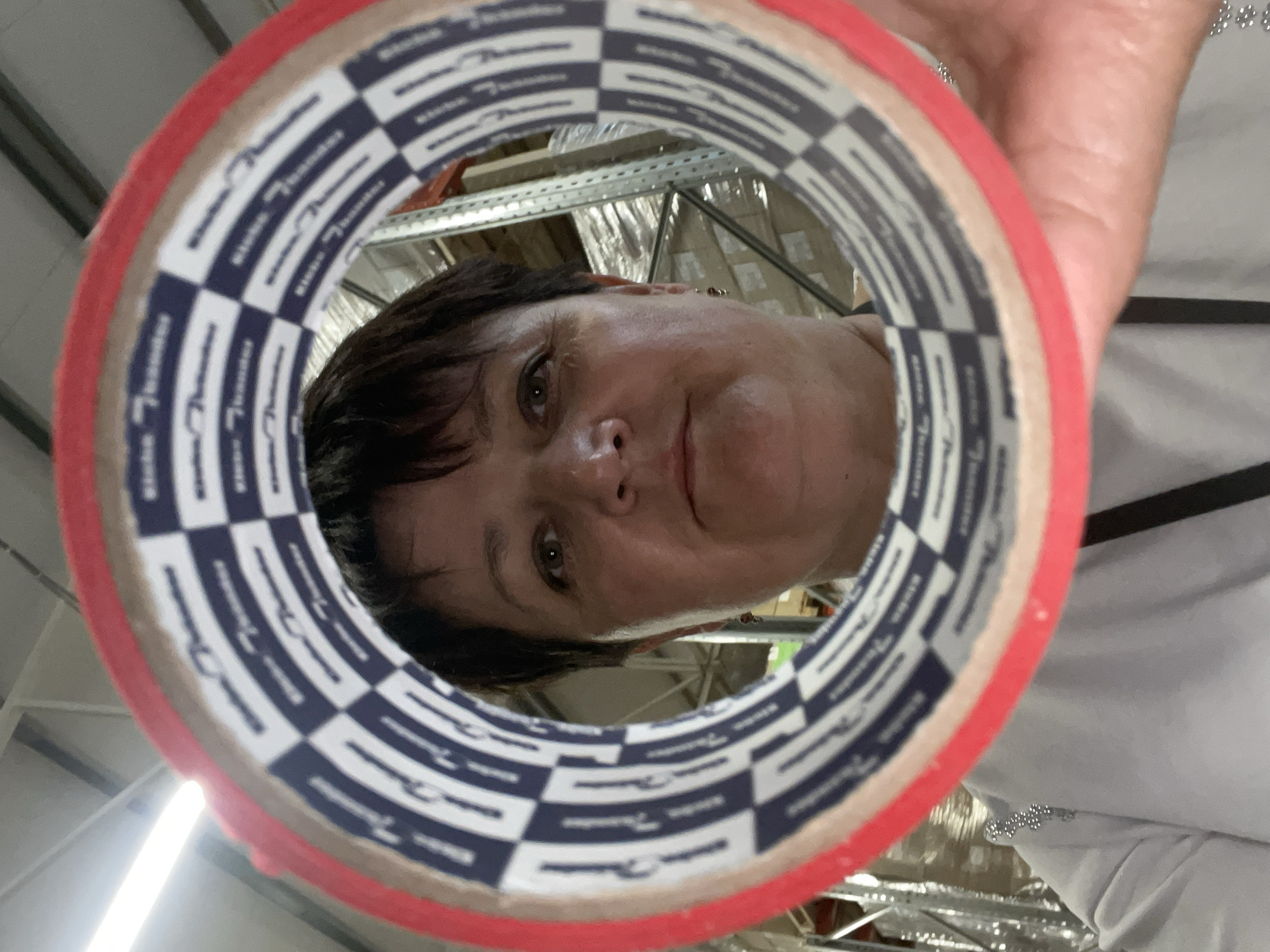 In a warehouse environment, a woman is holding a roll of red tape in front of her face, creating a framing effect. She has short, dark hair and is looking directly at the camera. The tape has a distinctive blue and white checkered pattern with repeated text. Behind her, metal shelves are visible, stocked with cardboard boxes. The lighting suggests overhead fluorescent lights, typical of an industrial setting. The location is identified as Терволово, Russia.