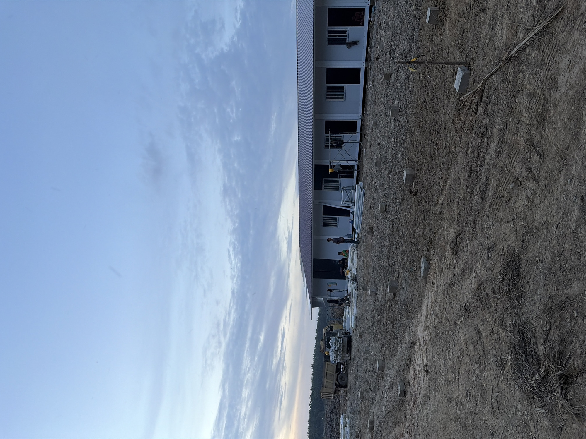 The image shows a construction site in Simpangempat, Indonesia. A long, two-story modular building is situated alongside a dirt lot. Construction workers are present near the building. A truck loaded with building materials is also visible. The sky is partly cloudy. The ground appears to be prepared for construction.