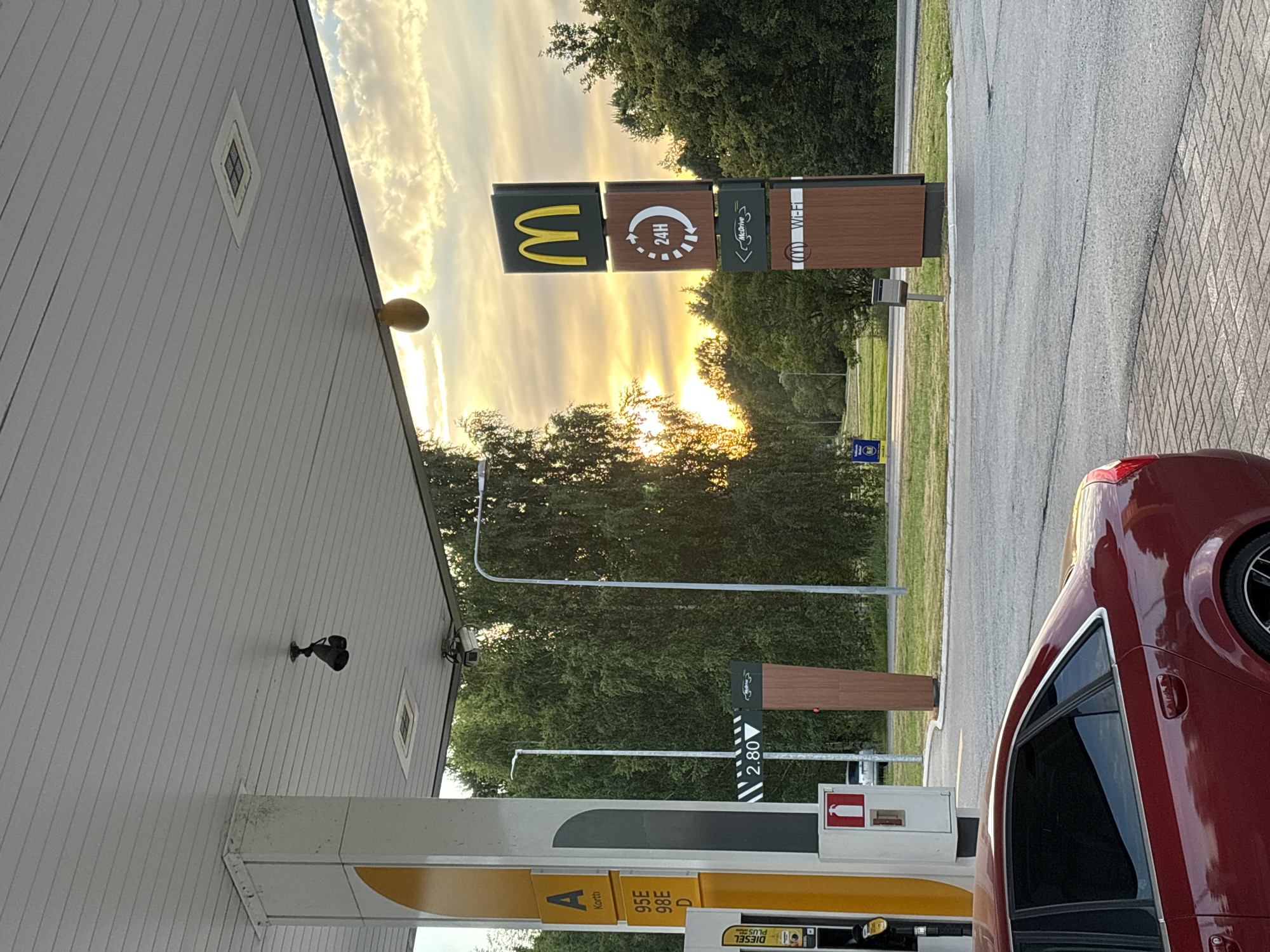 This image depicts an outdoor scene at a McDonald's restaurant, likely a drive-thru, in Pori, Finland. The time of day appears to be late afternoon or early evening, as indicated by the warm, golden light of the setting sun illuminating the sky and casting long shadows. The sky is filled with dramatic, streaky clouds.
The primary subjects are the McDonald's signage and a red car at a gas pump. The McDonald's sign is a prominent feature, featuring the iconic golden arches logo on a dark background. Adjacent to it are smaller signs indicating "24H" and "McDrive" as well as "Wi-Fi." The McDonald's building has a light-colored, corrugated exterior. Gas pumps are visible, with one partially obscured by the red car. The car is a dark red sedan, positioned at the pump.
The setting includes a paved area for vehicles, with a brick-patterned sidewalk or pathway. Beyond the immediate area, there is a line of green trees, suggesting a location with natural surroundings. A small blue sign with white text is visible in the distance, which might be a road sign or information board. The overall mood is serene, with the warm light creating a peaceful atmosphere.
Visible text includes "24H," "McDrive," "Wi-Fi," and what appears to be pricing information on the gas pump displays, such as "2.80" and labels for different fuel types like "DIESEL." There are also labels on the gas pump canopy, including "A" and what appears to be "Kortti." A small fire extinguisher cabinet is also visible. The image is taken from a slightly low angle, looking up towards the McDonald's sign and the sky.