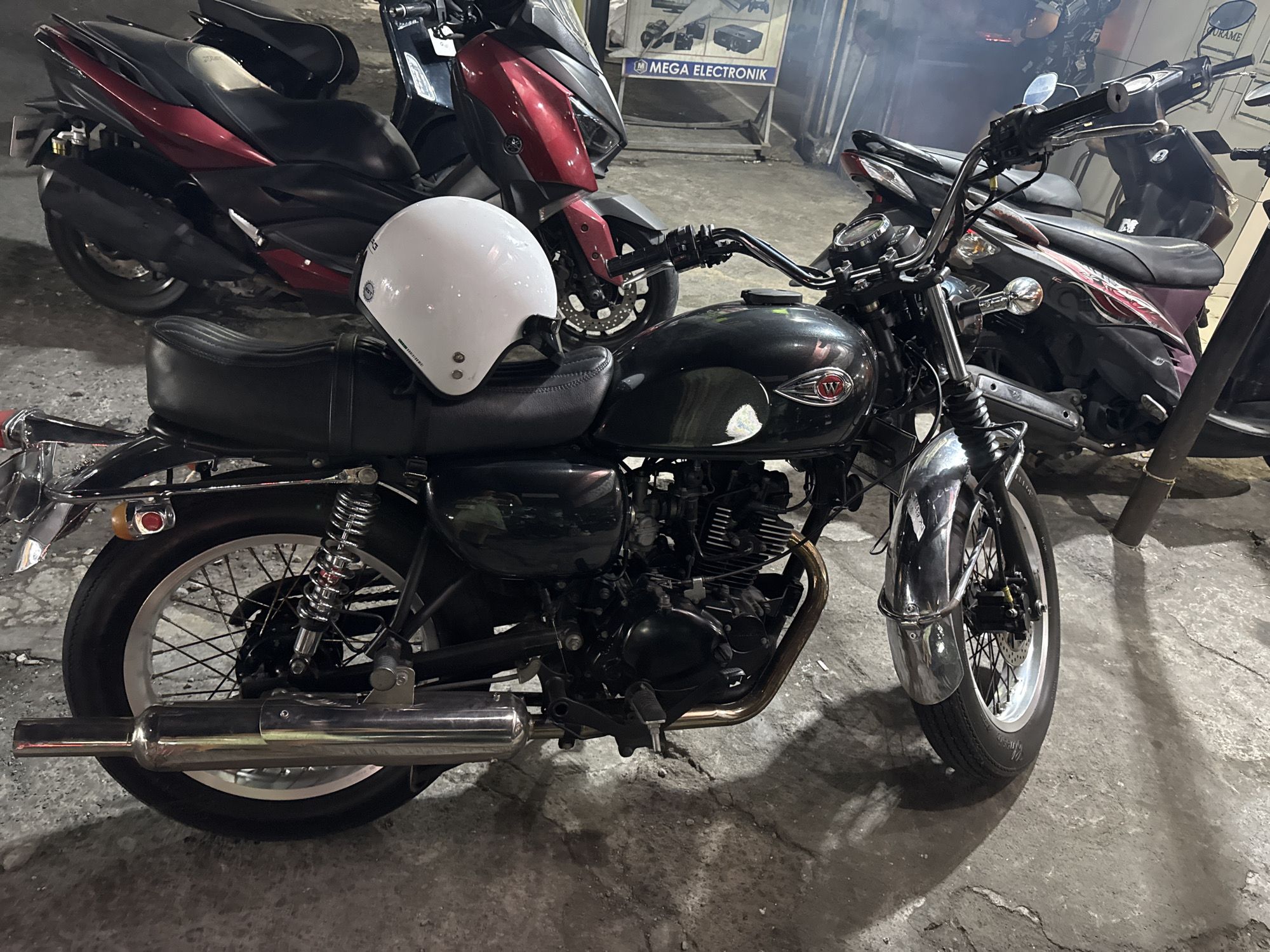 This media file captures a still shot of a black motorcycle parked on a concrete surface, likely outdoors at night. The motorcycle is the primary subject, with a white helmet placed on its seat. In the background, another red and black scooter-like motorcycle is visible, along with a sign that reads "MEGA ELEKTRONIK." The lighting suggests it's evening or night, with artificial light sources casting shadows on the ground. The scene appears to be a street or parking area in a commercial district, given the presence of the electronics store sign. There is a hint of smoke or mist in the air, adding a slightly atmospheric quality to the image. The motorcycles and the helmet are the main objects, with no people visible. The overall mood is neutral, focusing on the parked vehicles in their urban setting.