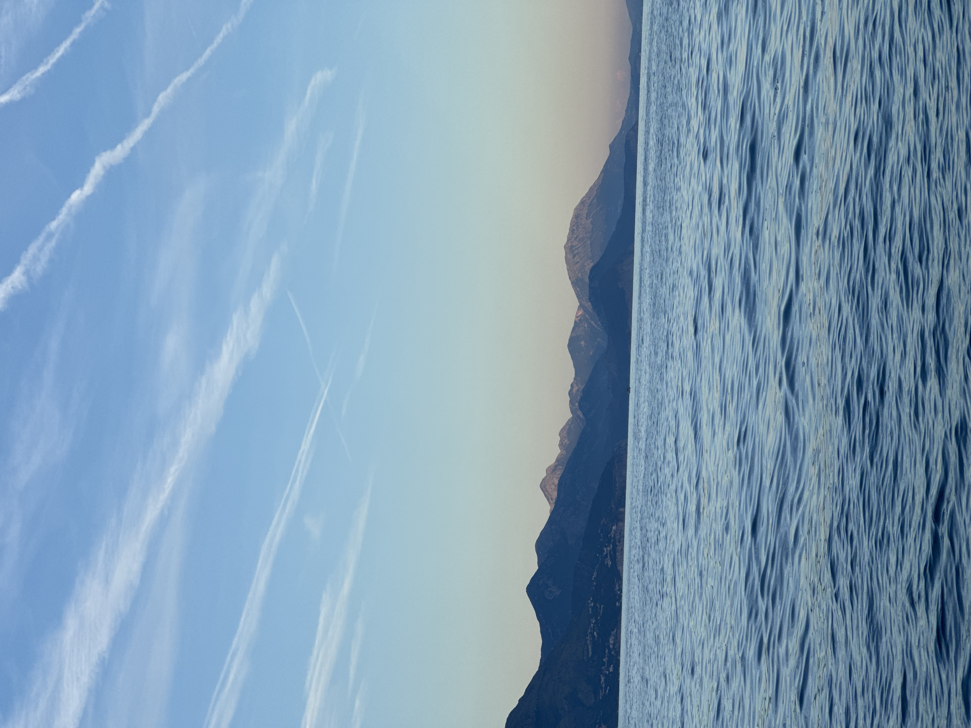 The image presents a serene landscape captured near Prijevor, Montenegro, featuring a body of water in the foreground, a mountain range in the middle ground, and a pale blue sky in the background.
The lower third of the image is occupied by calm, deep blue water, likely a sea or large lake, characterized by numerous small, parallel ripples and waves across its surface, indicating gentle movement. The color of the water subtly shifts, exhibiting lighter blue reflections in certain areas. No boats, people, or objects are visible directly on the water's surface in the immediate foreground.
The middle ground, extending horizontally across the frame, consists of a dark, forested mountain range. These mountains are predominantly dark blue-grey, appearing to be in shadow, with varied peaks and contours. Along the lower slopes of the mountains, particularly on the left side, faint visual cues suggest the presence of scattered structures or a small settlement, though specific architectural details are not discernible. The more distant peaks appear lighter and hazier due to atmospheric perspective.
The upper two-thirds of the image display a clear, pale blue sky. The sky transitions to a slightly lighter, almost white-blue tone near the horizon line. Several distinct white contrails from aircraft are visible, crisscrossing the sky in various diagonal patterns. Some contrails are sharply defined, while others are more diffused, suggesting varying ages or altitudes. The overall lighting is soft and diffused, indicating either early morning or late afternoon. No direct sunlight or harsh shadows are prominent.