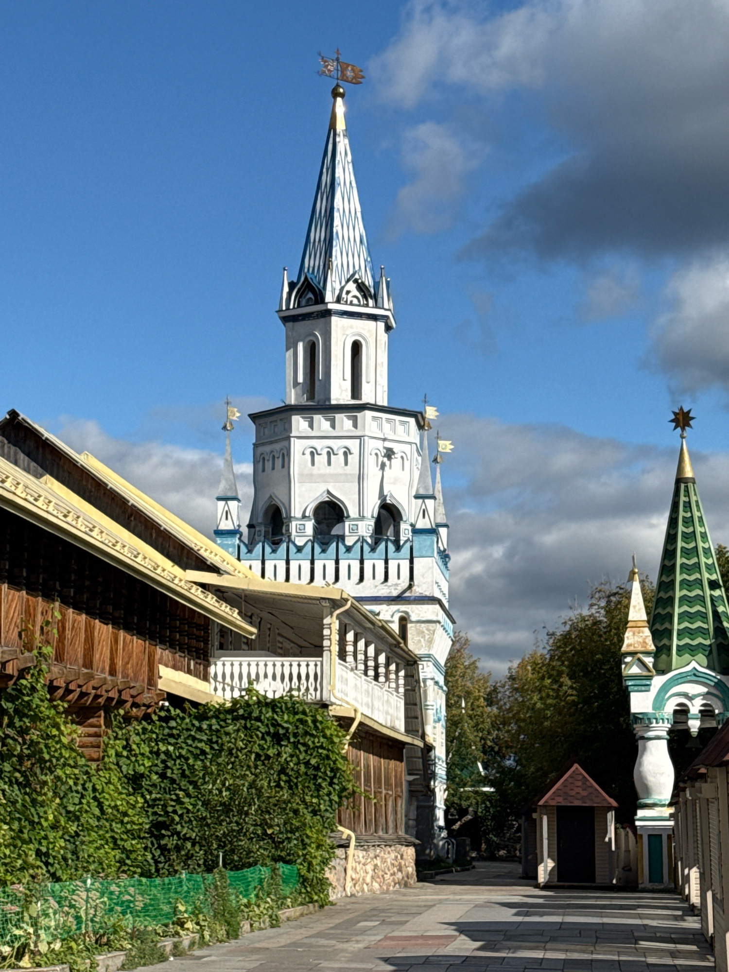 The image depicts a narrow, paved walkway leading through an area with traditional Russian architecture. On the left, a wooden building with a yellow-trimmed roof and a white balcony is covered in lush green ivy. To the right of the walkway, a smaller, dark-colored structure with a red roof stands. Further down the path, two prominent towers rise into a blue sky dotted with gray clouds. The taller tower on the left is white with blue trim and topped with a weather vane. To its right, a shorter, green-tiled spire is visible, crowned with a gold star. The overall scene suggests a historical or themed district, characteristic of areas like Kolomenskoye in Moscow, Russia, which features preserved historical buildings and architectural styles. No people are visible in the image.