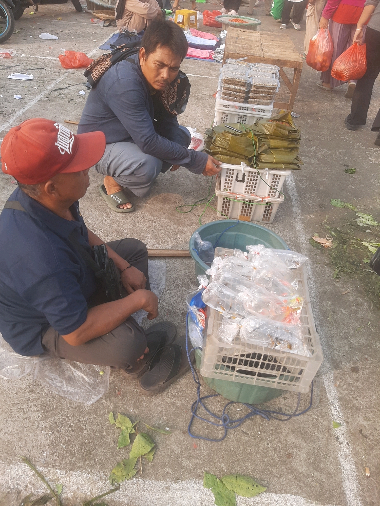 The image depicts an outdoor scene, possibly a street market, in Bekasi, Indonesia. Two men are the primary subjects. The man in the foreground, wearing a red baseball cap and a dark blue polo shirt, is seated on the ground. He has a black bag slung across his chest and appears to be looking downwards or towards the man behind him. The second man, wearing a dark blue long-sleeved shirt and light grey pants, is crouched behind the first. He is looking down at a stack of green-wrapped food items.
The environment includes a concrete ground with scattered debris, including leaves and plastic. Several objects are present: a wooden stall or table behind the men with stacks of packaged food items on it, white plastic crates filled with wrapped food and clear plastic bags containing what appear to be fish or aquatic creatures, and a large green basin. In the background, other individuals and market stalls are visible, suggesting a busy public space. The lighting indicates it is daytime.
