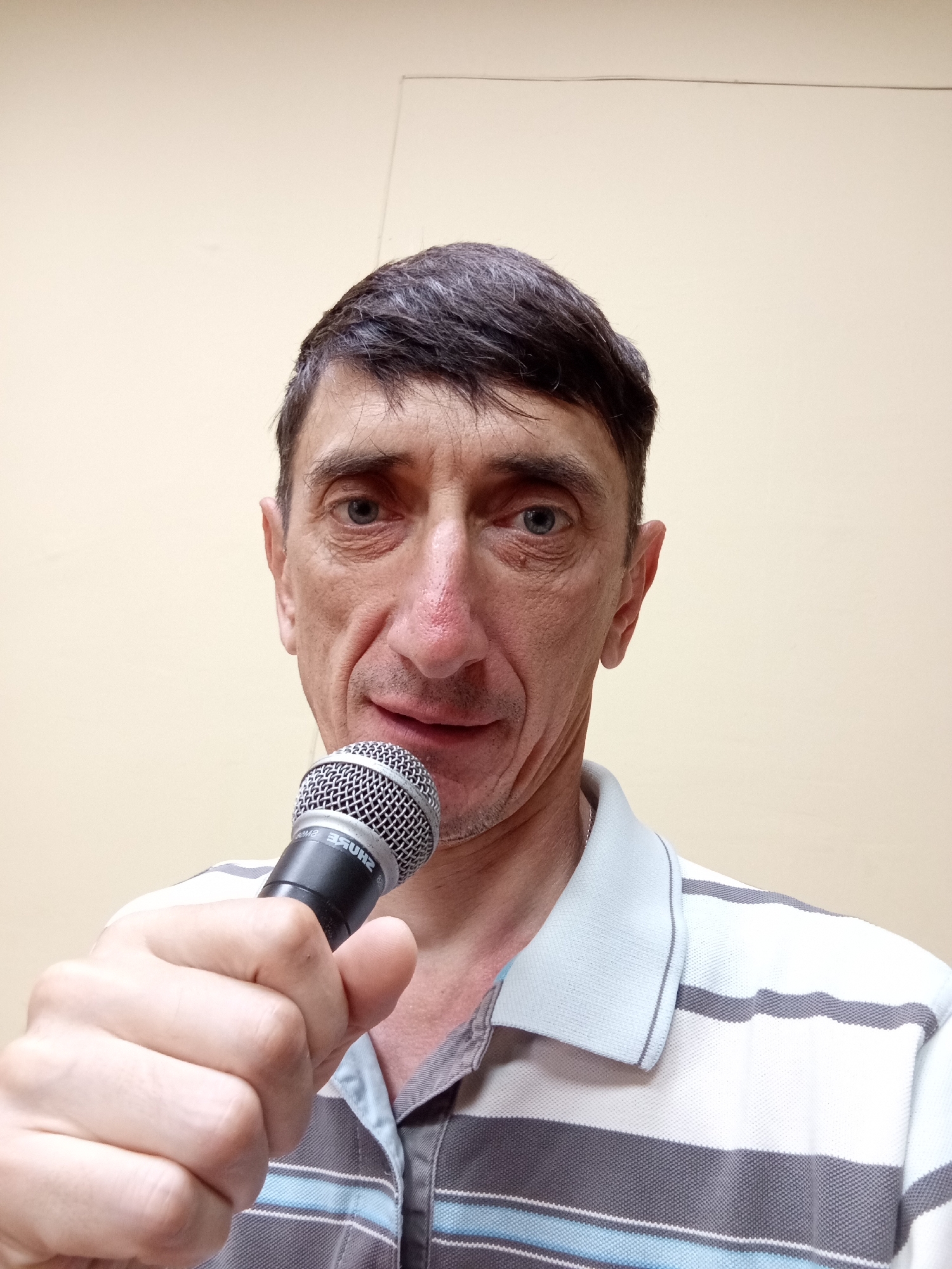 An over-the-shoulder, medium shot captures a man holding a microphone near his face against a plain, beige wall. The man, with short, dark hair and weathered skin, gazes directly at the camera. He wears a horizontally striped polo shirt in shades of white, gray, and blue. His right hand grips the microphone, which is positioned just below his mouth. The background is a simple, unadorned wall with a faint horizontal line visible near the top. The lighting is even and appears to be artificial. The scene is devoid of any specific identifying markers of location within Крыўлянскі сельскі Савет, Belarus, offering no visual clues to its specific setting.