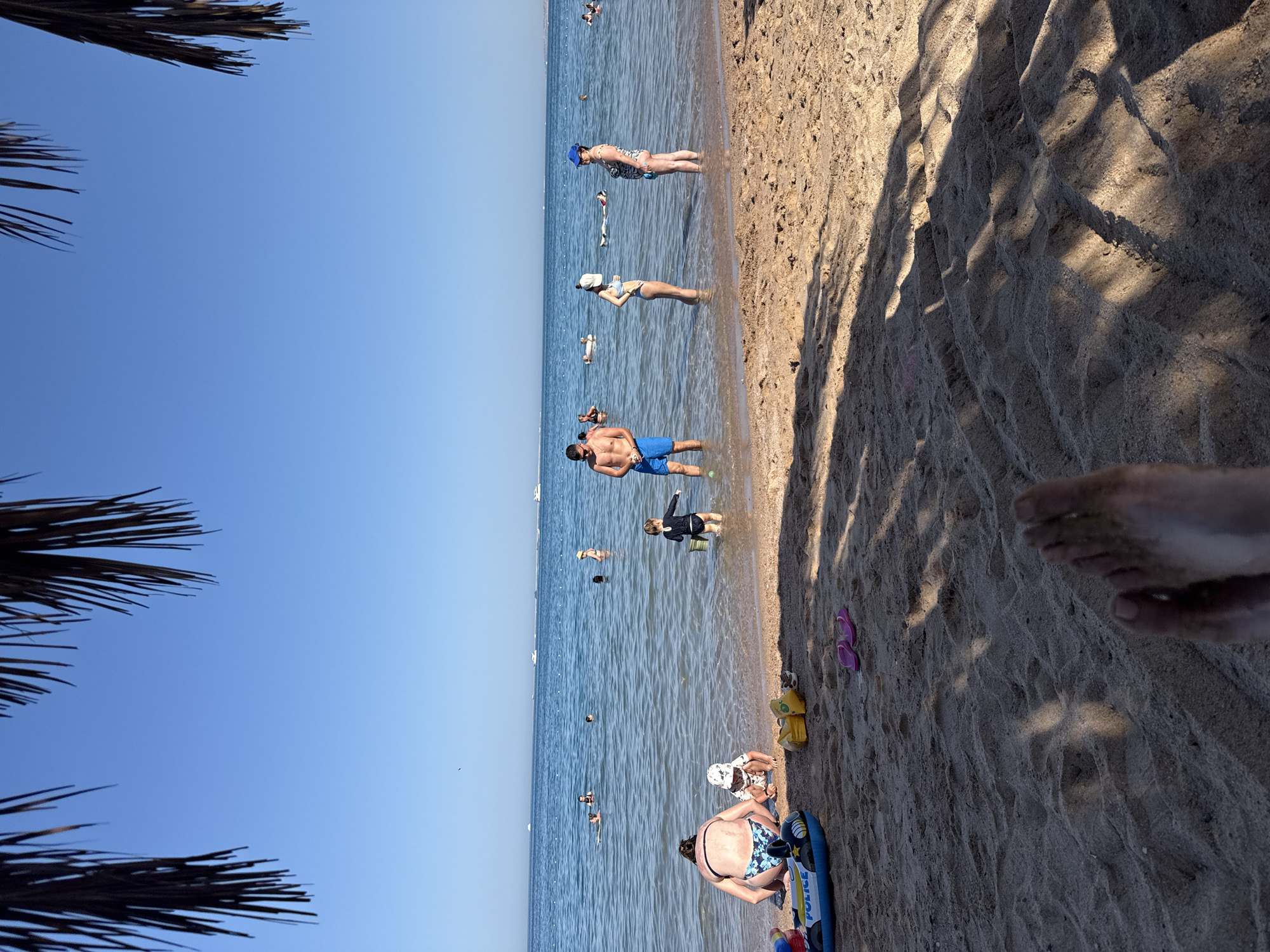 The image depicts a sunny day at a beach in Hurghada, Egypt. The scene is primarily composed of sand and the calm, blue sea. Several people are visible enjoying the water and the beach. In the water, some individuals are wading, while others are swimming or floating. On the sandy shore, people are seen relaxing, with some children playing.
Notable details include the clear blue sky and the presence of palm fronds in the foreground, suggesting a tropical or subtropical environment. The lighting indicates it is daytime, likely late morning or early afternoon, given the shadows cast by the palm fronds and the bright sunlight. The overall mood appears relaxed and leisurely, characteristic of a beach vacation.
Objects present include a yellow inflatable toy and pink flip-flops on the sand. A blue inflatable toy with the word "POLICE" printed on it is also visible. There are no clear indications of specific emotions, but the activities suggest enjoyment and relaxation. The location is specified as Hurghada, Egypt, which aligns with the visual cues of a sandy beach and clear blue water.