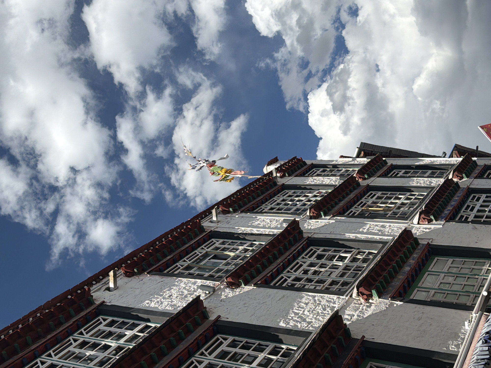 The image presents an upward-looking view of a multi-storied traditional building, likely an ancient monastery or palace, set against a bright blue sky filled with large, white cumulus clouds. The building, consistent with Tibetan architectural styles and located in Chengguan District, China, features a light-colored facade with numerous rows of small, dark-framed windows. Each window has multiple panes, and many are covered by decorative, red-colored awnings or eaves that add a striking contrast to the white walls.
Long strings of colorful prayer flags, predominantly yellow, green, and white, are visible in the upper left portion of the frame. They are suspended from the building's upper reaches and are actively fluttering in the wind against the backdrop of the sky. The bright sunlight suggests it is daytime, casting clear shadows on the building's textured walls and highlighting its architectural details.
In the far upper right corner, a small, cropped section of a red flag with a yellow star is visible, indicating the flag of China. No people are visible in the scene, and the activity is limited to the wind blowing the prayer flags. The overall impression is one of cultural significance and serenity under a vast, open sky. No other discernible text is visible in the image.