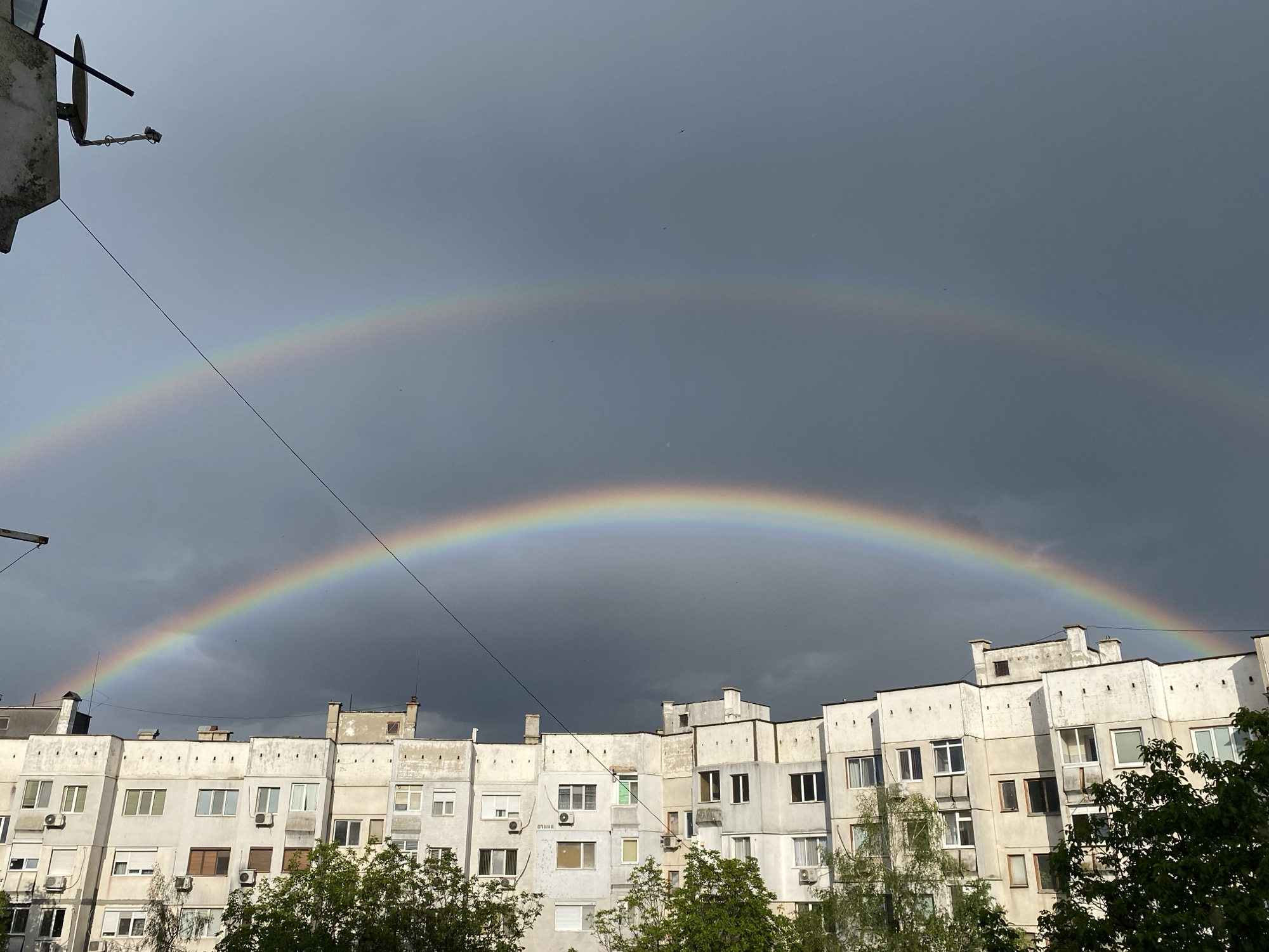 The image depicts a double rainbow arched over a city landscape. The lower, brighter rainbow is more prominent, spanning across the tops of several similar-looking, multi-story apartment buildings. The buildings are of a pale color with numerous windows and what appear to be air conditioning units attached to some facades. Trees with green foliage are visible at the base of the image, partially obscuring the lower portions of the buildings. Above the lower rainbow is a fainter, secondary rainbow. The sky is predominantly gray and overcast, suggesting a recent or ongoing rain event. The image was likely taken in the city of Pleven, Bulgaria, considering the request. A satellite dish and wires are visible on the top left of the frame. There are no people visible in the image.