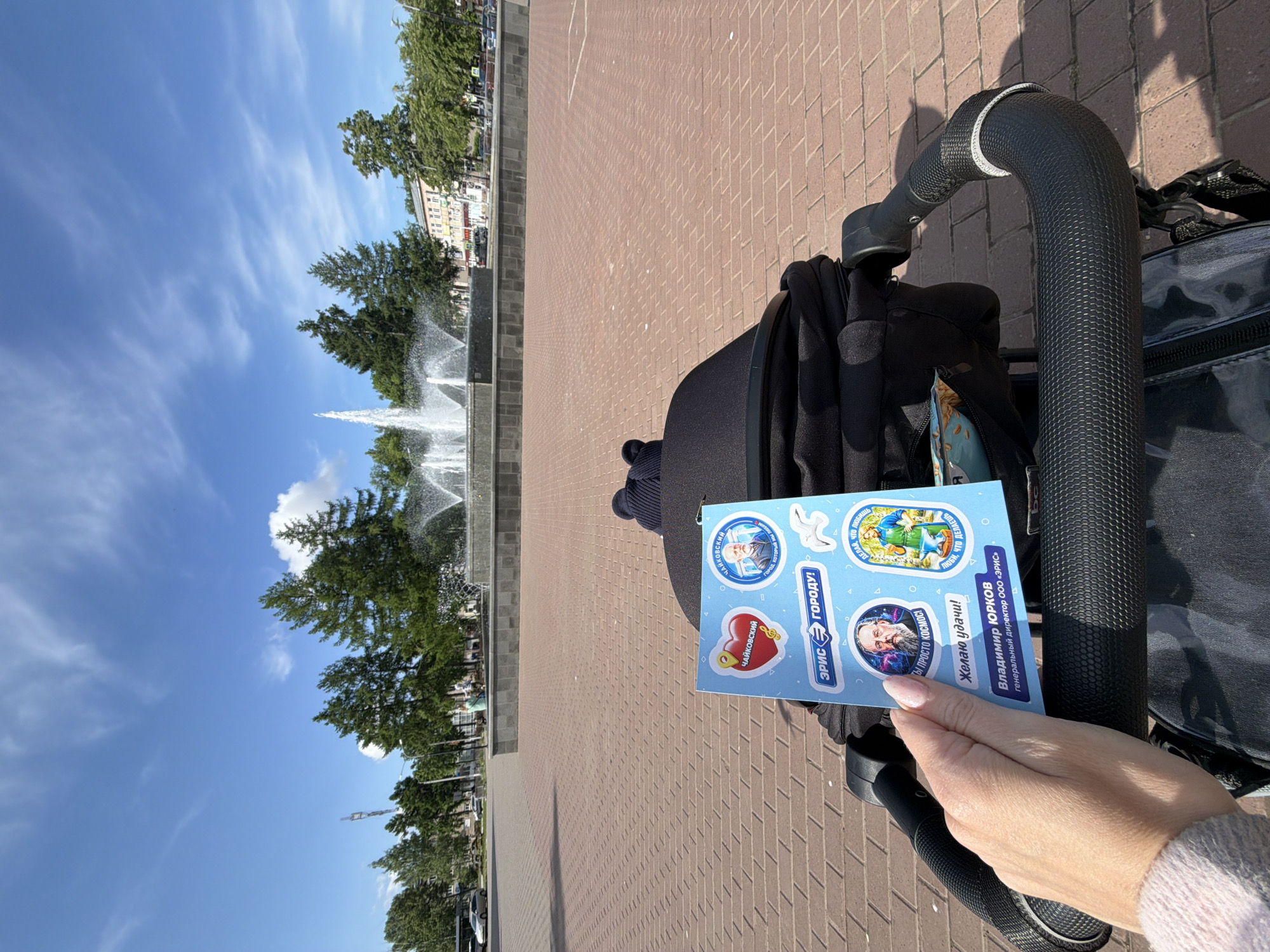 The image shows a person holding a sticker sheet promoting Vladimir Yurkov, general director of “ERIS” and featuring images of Pyotr Ilyich Tchaikovsky in the city of Chaikovsky, Russia. The sheet also includes phrases like "Like Tchaikovsky," "The city that we love," and "Do what you love." The photo appears to be taken from the perspective of someone pushing a black baby stroller on a brick pedestrian area with a large fountain in the background. Trees line the edge of the square behind the fountain. The sky is blue with scattered clouds.