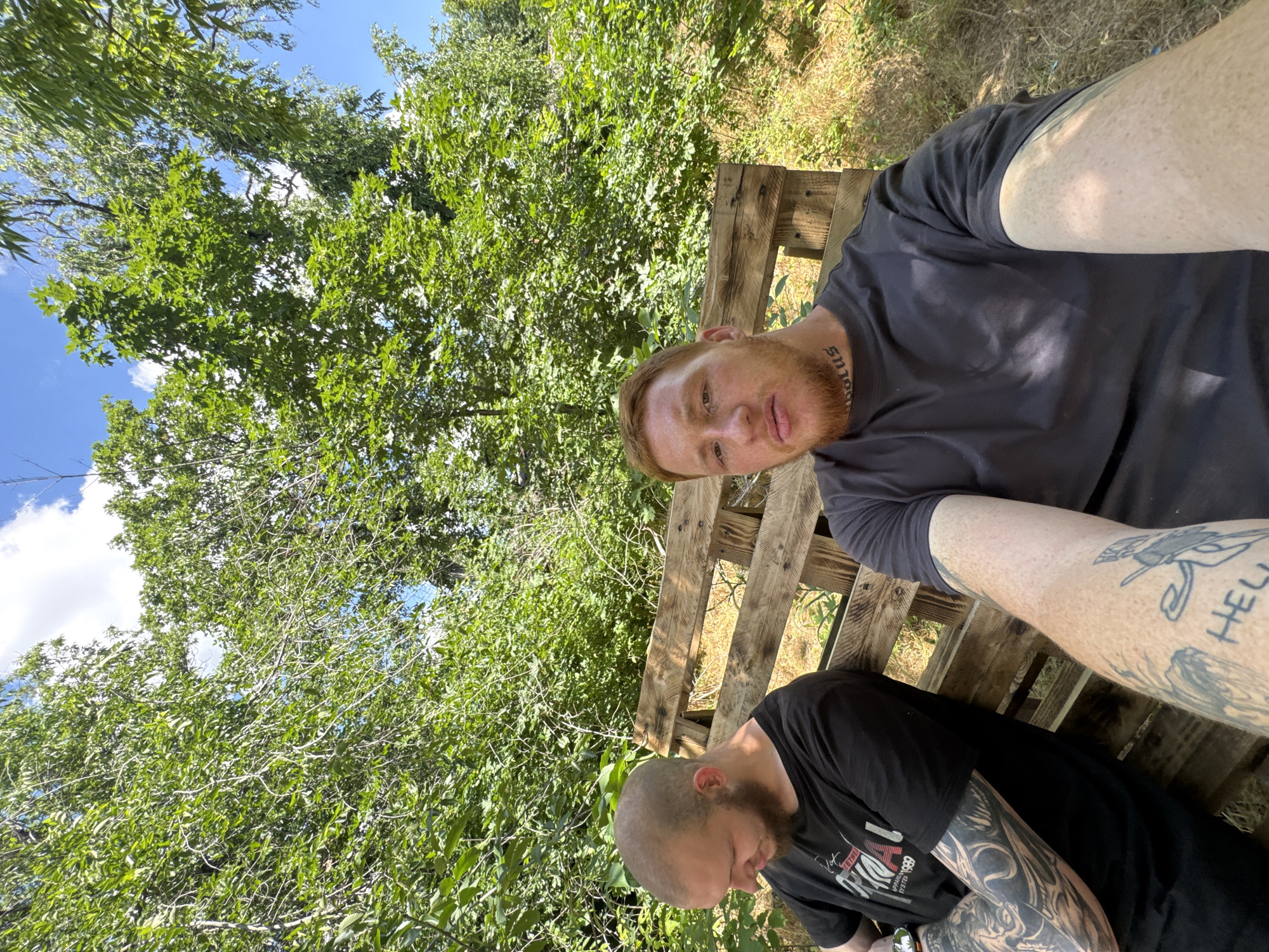 Two men are sitting on a wooden bench outdoors. The man on the left is bald with a beard and is wearing a black t-shirt with a graphic print. He has a heavily tattooed left arm. The man on the right has red hair and a beard. He is wearing a dark grey t-shirt with a tattoo on his neck that reads "kolos". His right arm is also tattooed. The bench is made of rough wooden planks. The background is filled with lush green foliage and trees under a bright blue sky with scattered clouds. The ground around the bench is covered in dry grass and vegetation. The lighting suggests it is a sunny day.