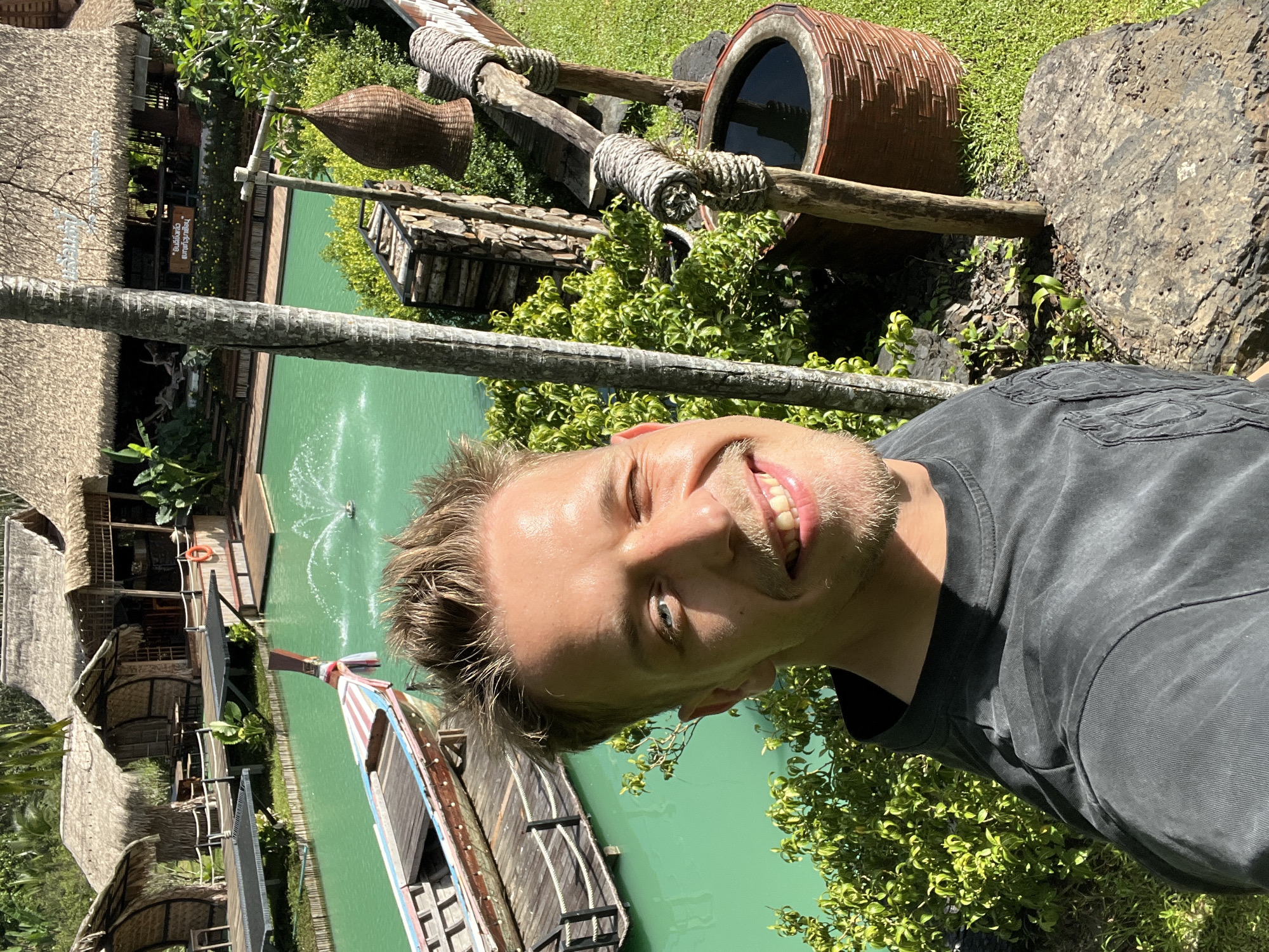 The media file is a portrait-oriented selfie taken outdoors in a tropical setting. The subject is a man with short blonde hair and a beard, smiling at the camera. He is wearing a dark grey t-shirt. The background suggests a resort or tourist attraction with thatched-roof buildings and a vibrant green body of water, possibly a pool or a canal. A traditional wooden boat with colorful stripes is visible on the water.
In the upper right of the frame, there is a large, rustic clay pot with a dark interior, placed on a wooden structure. Woven baskets hang from wooden beams, and there are stacks of wood. Lush green foliage surrounds the area. The sunlight suggests it is daytime, and the weather appears to be sunny and warm. The location is identified as Tambon Pa Kae Bo Hin, Thailand, which aligns with the visual cues of tropical vegetation and traditional architecture. A small fountain is visible in the water, adding to the scenic ambiance. There is a red life preserver hanging on a railing near one of the buildings. Text is visible on a sign in the upper left portion of the image, but it is difficult to read clearly. The overall mood is relaxed and happy, reflecting the subject's smile and the pleasant environment.