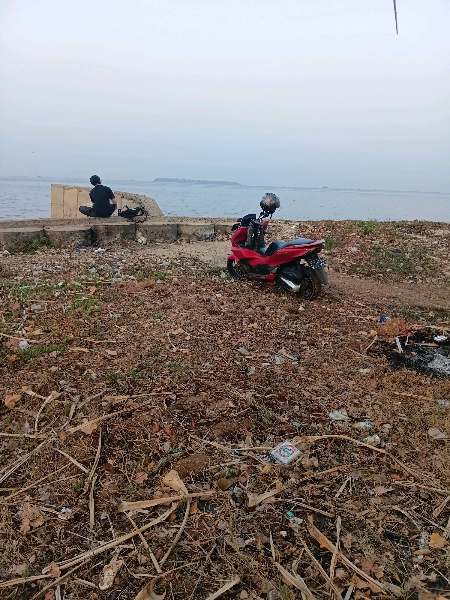 A person sits facing a body of water at a concrete barrier, with a red scooter parked on the ground behind them. The ground is covered in dry grass and debris. The sky is overcast. The location is identified as Cikal, Indonesia.