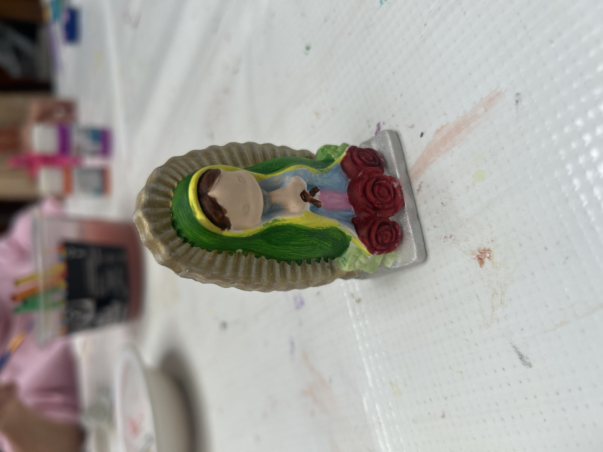 A figurine of the Virgin of Guadalupe lies on a white, textured surface. The figure is predominantly light tan with accents of green, gold, red, and blue. The setting is likely indoors, but the specific location is unknown.