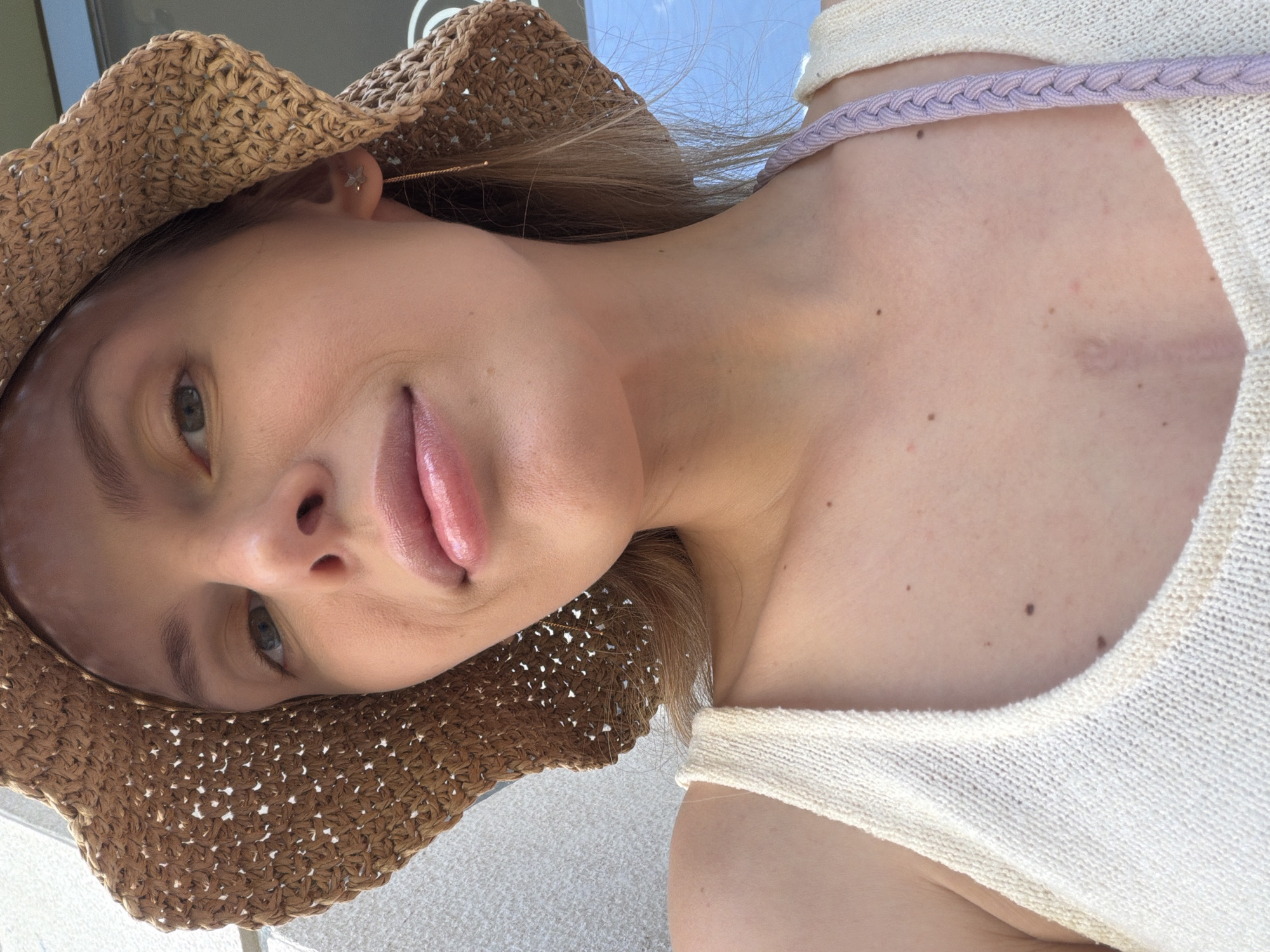A woman wearing a straw hat and a light-colored top with a braided strap is posing for a selfie. The setting appears to be outdoors, possibly near a light-colored surface like a sidewalk or patio. The sky is visible in the background. A possible scar is noted on the upper chest. The location of Budva, Montenegro, cannot be identified from the image.