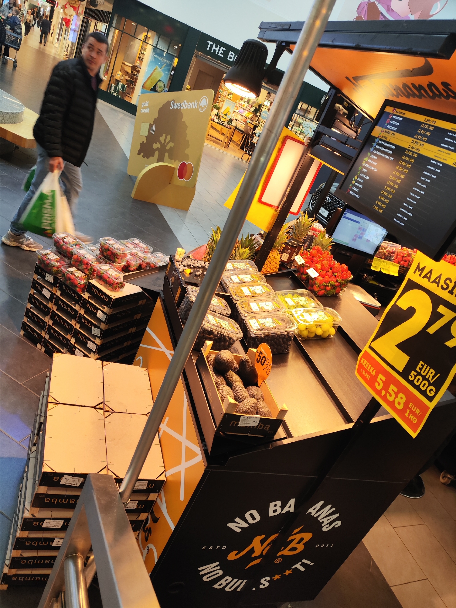 The image depicts a fruit stand within a shopping mall, possibly in Tallinn, Estonia, given the "Swedbank" branding, a prominent bank in the Baltic region. A man, dressed in a black puffer jacket and grey jeans, walks towards the left, carrying two shopping bags, one green and one white. The fruit stand displays various fruits in plastic containers, including strawberries, grapes, blueberries, and avocados. Prominent signage features prices in EUR and indicates fruit types such as "MAASIKAS" (strawberry) and "KREEKA" (grape). A digital screen displays more product information. The overall environment suggests an indoor shopping center with a tiled floor and illuminated storefronts in the background.