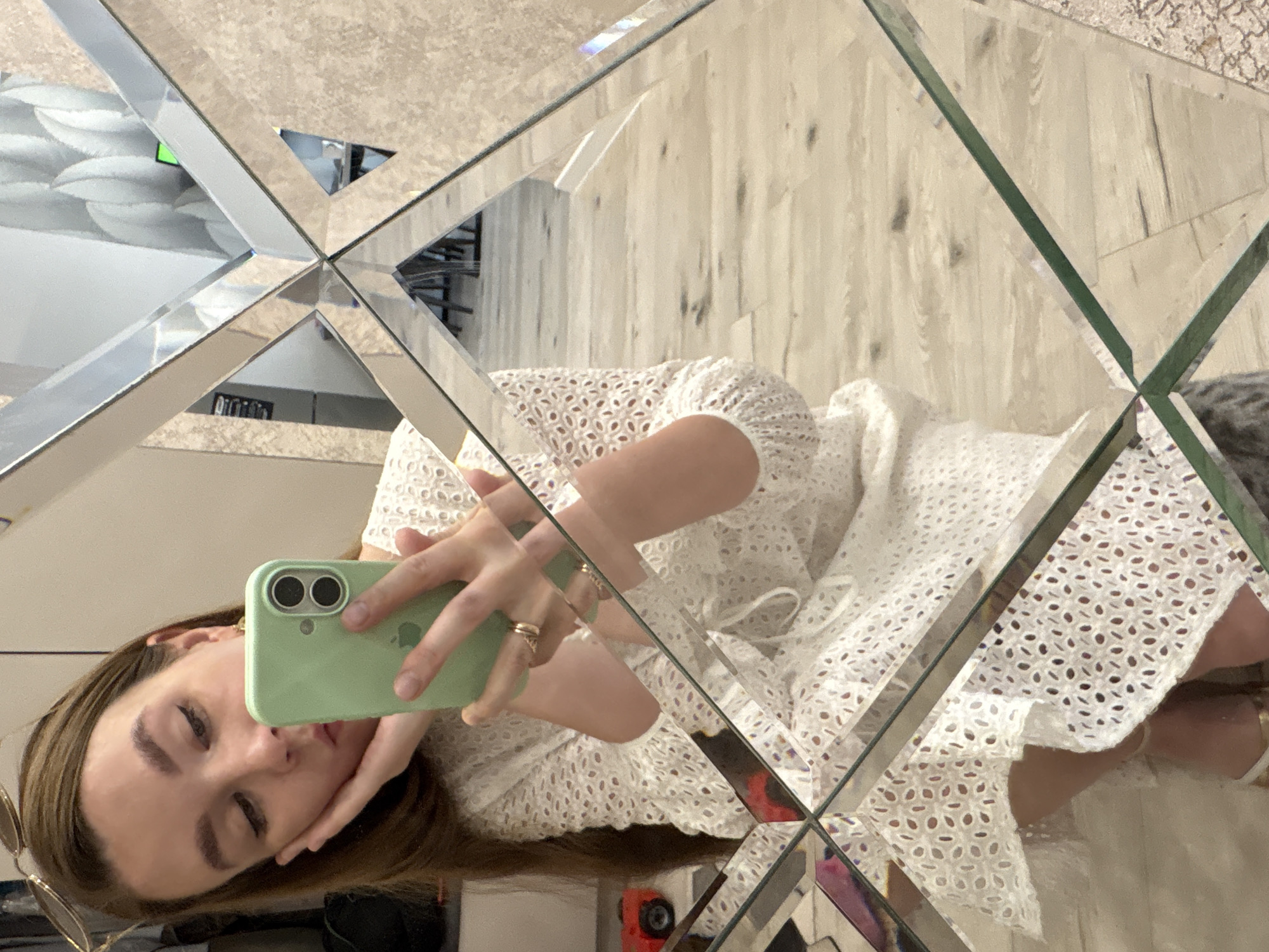 A young woman with long brown hair takes a mirror selfie in what appears to be a residential interior. She is wearing a white eyelet dress and gold strappy sandals. She holds a light green smartphone in her left hand, which is positioned to capture her reflection. Her right hand is resting on her cheek, and she is looking down at her phone. Sunglasses are perched on top of her head. The mirror is framed by geometric glass panels, creating a fragmented reflection of the room, which includes a light-colored wooden floor and textured walls. A small red toy car is visible in the reflection on the left side of the image. The scene is indoors and appears to be in a domestic setting. It is impossible to determine the specific location in Izhevsk, Russia, based on the visual information provided.