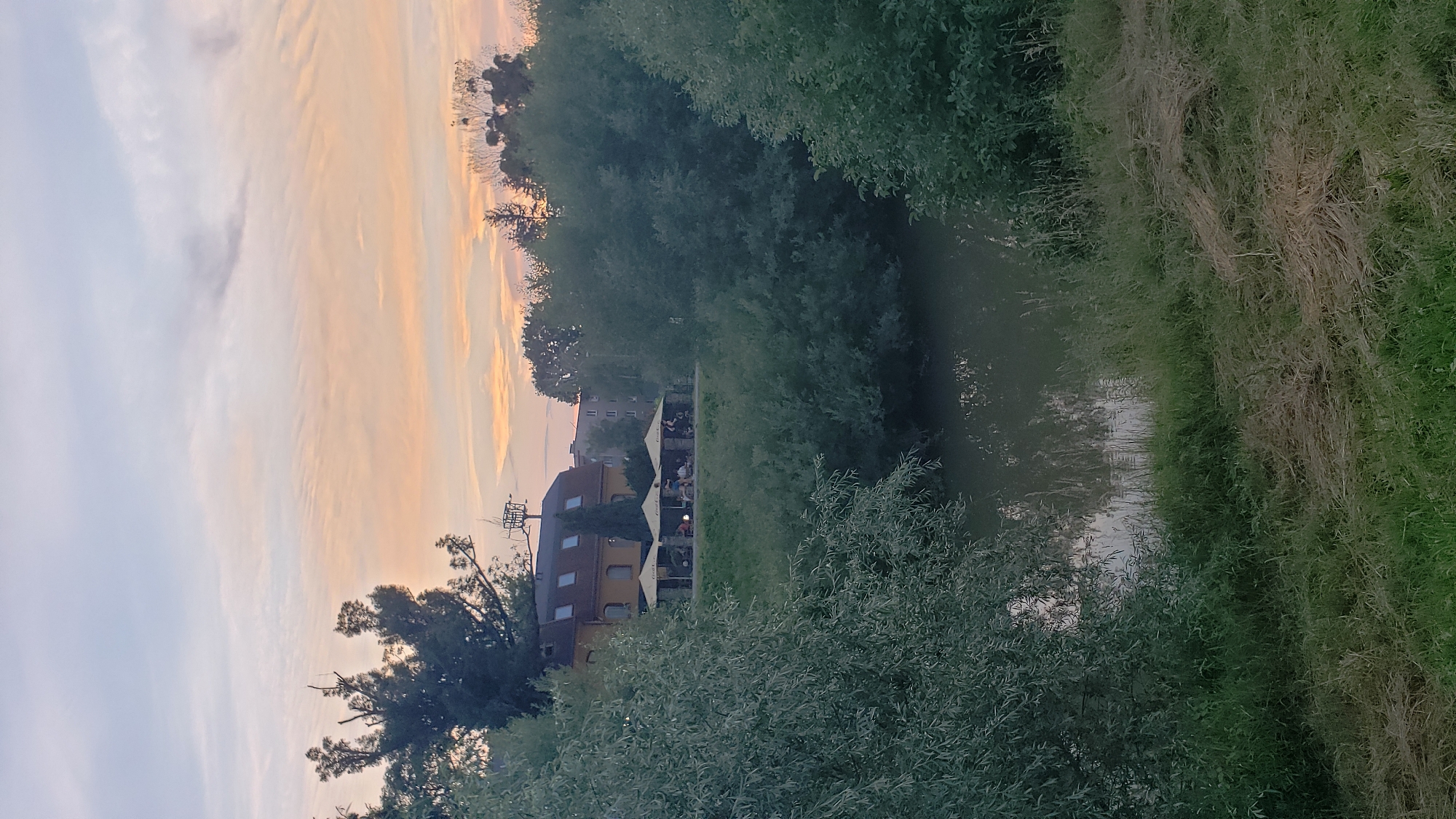 The image depicts a serene evening scene along a riverbank in Prešov, Slovakia. A building with a yellow facade and multiple dormer windows is visible on the elevated bank, with several yellow umbrellas providing shade over outdoor seating areas. People are seated at tables beneath the umbrellas, suggesting a cafe or restaurant setting. The river in the foreground reflects the soft colors of the sunset sky, which is a blend of pale blue, pink, and orange hues, with wispy clouds. Lush green trees and foliage line the river and surround the building, creating a natural and tranquil atmosphere. The overall impression is one of a peaceful, end-of-day moment in an urban natural setting.