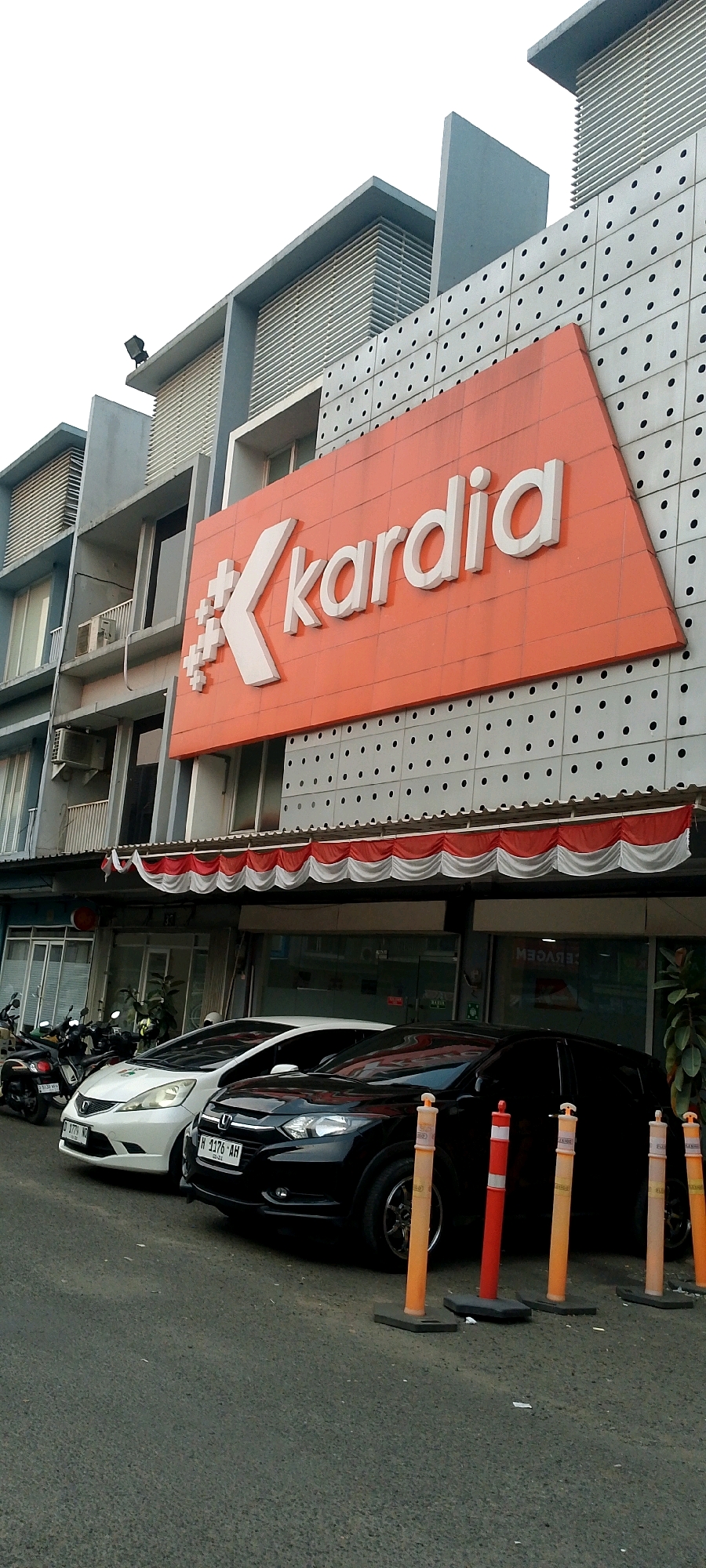 A low-angle, full-frame shot shows the exterior of a multi-story building with a large, orange sign featuring the white text "Kardia" and a stylized "K" symbol on the facade. The building has a modern architectural style with grey and white paneling, accented by horizontal window louvers and black circular perforations. Below the sign, a red and white striped awning is visible, beneath which are glass-fronted shop entrances. In the foreground, a paved street displays two parked cars: a white hatchback on the left and a black SUV on the right. Several orange and red traffic cones are placed in a line in front of the black SUV. Motorcycles are parked to the far left of the frame, near the building's entrance. The scene is outdoors, likely in an urban environment, given the presence of buildings, cars, and street paving. The lighting suggests it is daytime, possibly late afternoon or early morning, with overcast conditions. There are no people clearly visible in the image.