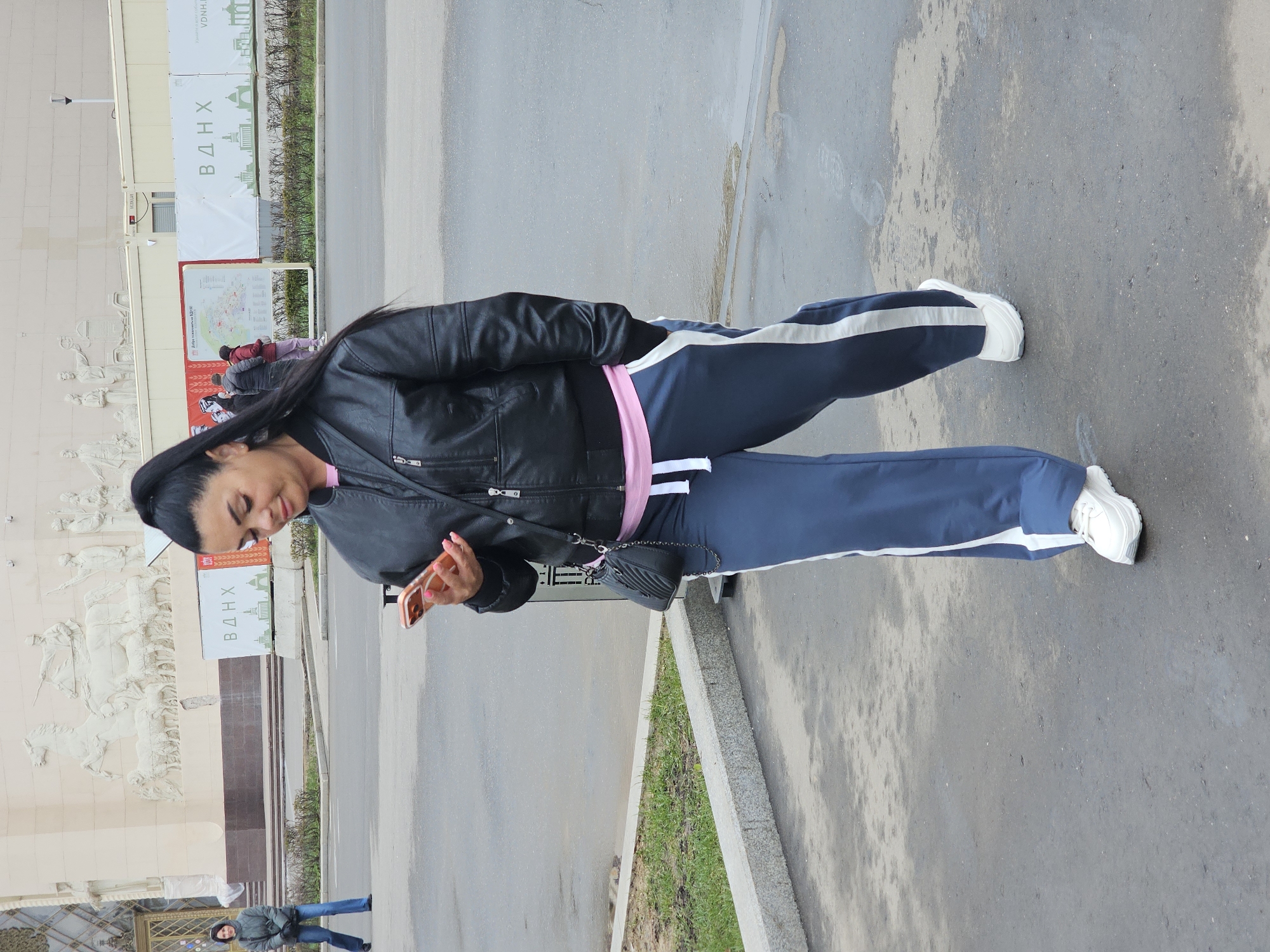 An outdoor, slightly tilted, medium shot captures a woman standing on a paved surface, looking down at a smartphone in her hands. She wears a black leather jacket over a pink top, navy blue track pants with white stripes down the sides, and white sneakers. A small black shoulder bag hangs across her body. Her dark hair is pulled back, and she exhibits a gentle, smiling expression.
In the background, a large, light-colored building with intricate bas-reliefs depicting figures and horses is visible, characteristic of Soviet monumental architecture. Adjacent to the building, a large white banner prominently displays the Cyrillic letters "ВДНХ" (VDNH), alongside a stylized building silhouette, identifying the location as the Exhibition of Achievements of National Economy in Moscow, Russia. Another sign, partially obscured, appears to be a map. The foreground consists of asphalt or concrete pavement, with a curb and a strip of green grass separating it from what appears to be a road or path. The lighting suggests it is daytime, with soft, diffused light indicative of an overcast sky. Other distant, blurred figures can be seen in the background.