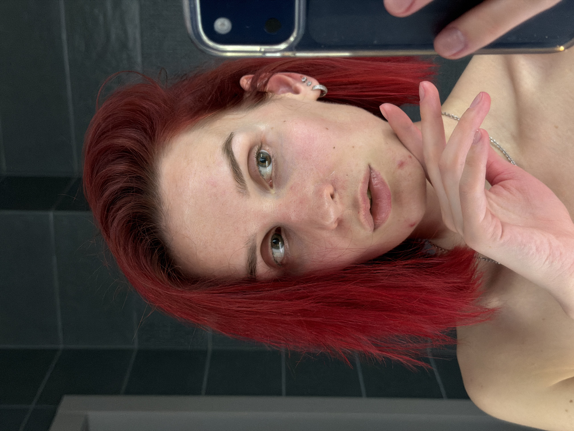 A woman with red hair is taking a selfie. She is holding a phone, the top portion of which is visible in the upper portion of the frame. She is wearing a small silver necklace. She has piercings in her left ear. The background appears to be a bathroom with gray tile. The location in Chaikovsky, Russia is unknown.