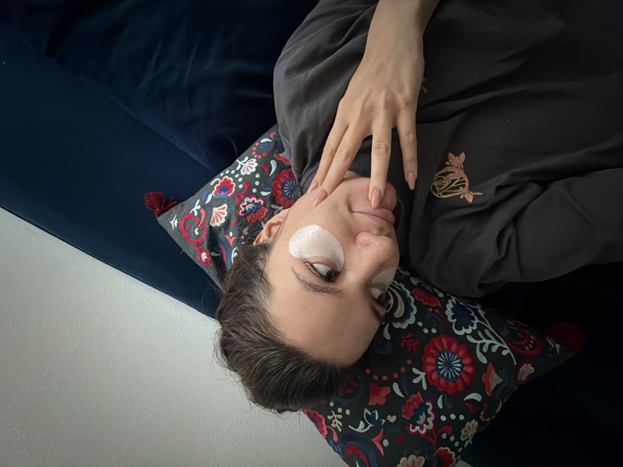 A woman with fair skin is lying on a patterned pillow, with a light-colored eye mask under her left eye. She is wearing a gray long-sleeved shirt with a design on the chest and has long, light pink painted nails on the hand resting on her face. The pillow is on a dark blue couch or bed. The background includes a white wall. It is impossible to tell if this photo was taken in Belgrade, Serbia.