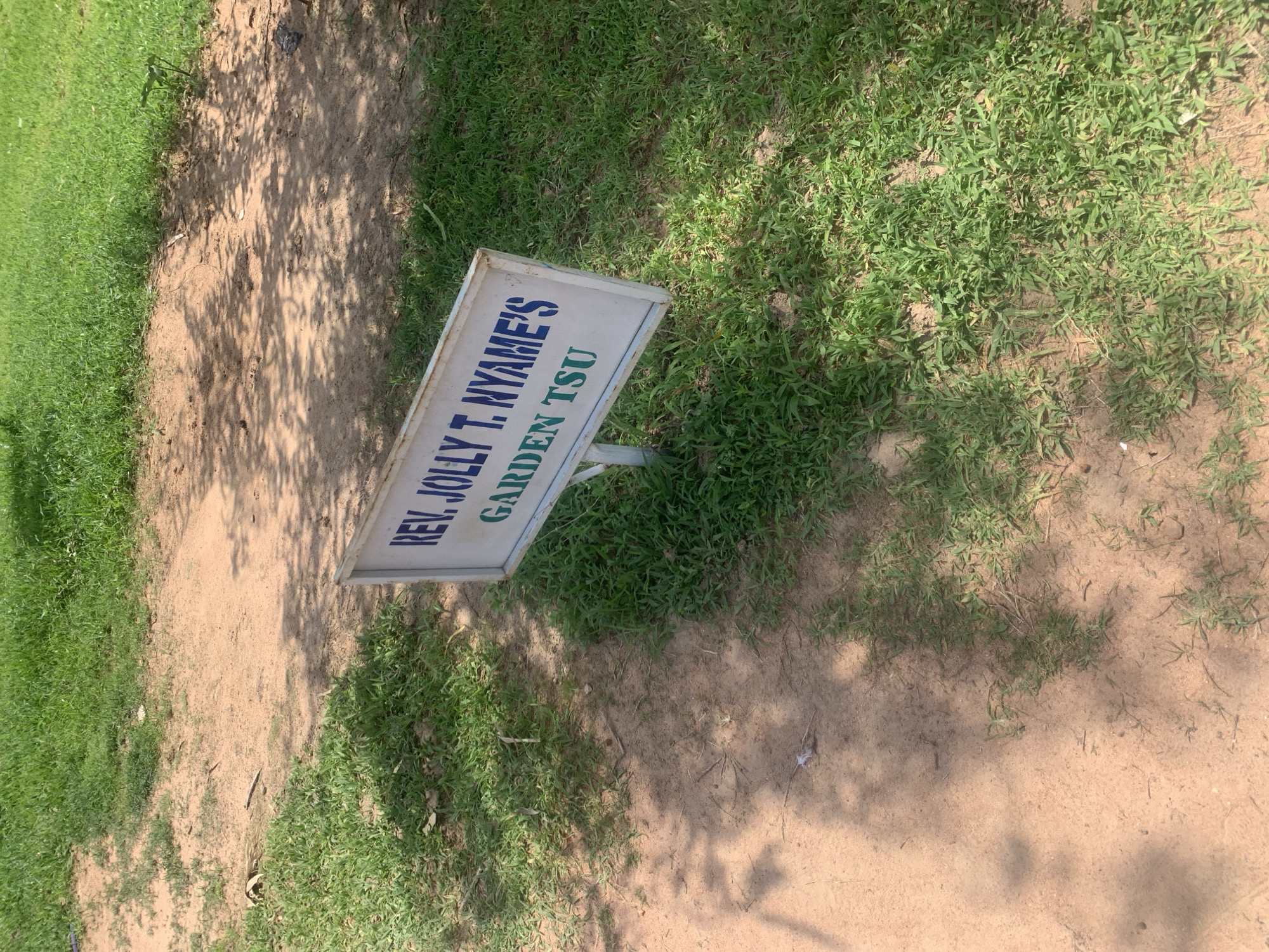 A white sign with a metal frame is placed in a grassy area with sandy patches. The sign has text written on it. The text in blue reads "REV. JOLLY T. NYAME'S" and below that, in green, it reads "GARDEN TSU". The sign is supported by a metal pole. The setting appears to be outdoors, with sunlight casting shadows from unseen trees onto the ground. The grass is green and appears to be dry in some areas. The time of day seems to be daytime, possibly mid-morning or afternoon, given the strong shadows. There are no people visible in the image. The overall scene is a simple outdoor setting with a sign marking a specific location.