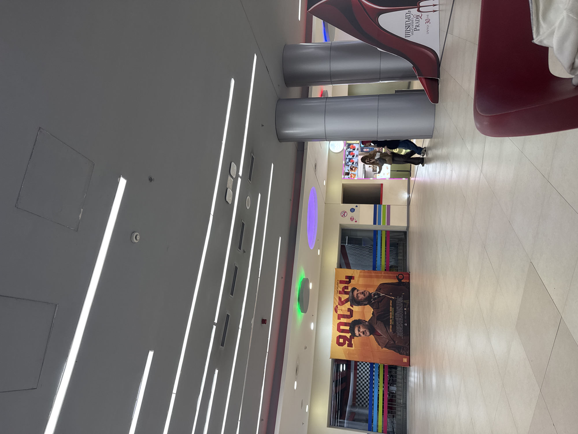 The image captures an interior space, potentially a shopping mall or entertainment complex in Yerevan, Armenia. Notable architectural features include large, cylindrical pillars and linear lighting fixtures embedded in the ceiling. A movie poster featuring two men, oriented vertically with Armenian text, is visible near an entrance. Two individuals are walking in the background, possibly interacting with a storefront. In the foreground, a red chair with what appears to be a white cloth draped over it sits adjacent to a display advertising a luxury shoe. The flooring is a light-colored tile, contributing to the overall bright and spacious environment.