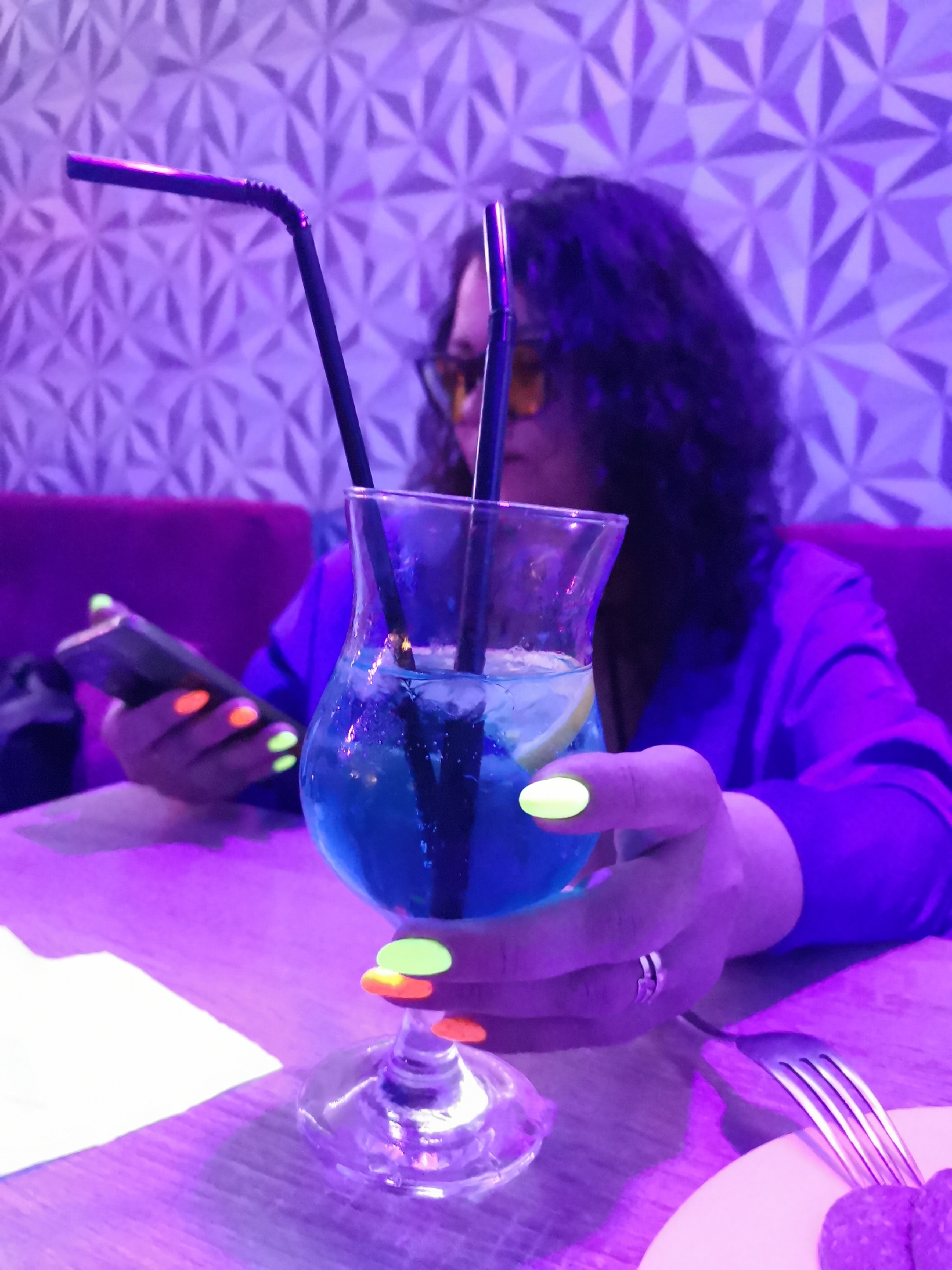 The image captures an indoor scene with a woman in the background holding a smartphone, while the focus is on a blue cocktail in the foreground. The woman has dark, curly hair and is wearing yellow-tinted glasses and a blue shirt. Her nails are painted with alternating neon green and orange colors. She is holding the smartphone in her left hand and a cocktail glass with her right. The cocktail has two black straws and a slice of lemon inside. The table is set with a fork and a plate, suggesting a restaurant or bar setting. The background features a geometrically patterned wall and a magenta-colored cushioned seating. The overall lighting is purple. The location appears to be a bar in an unknown city.