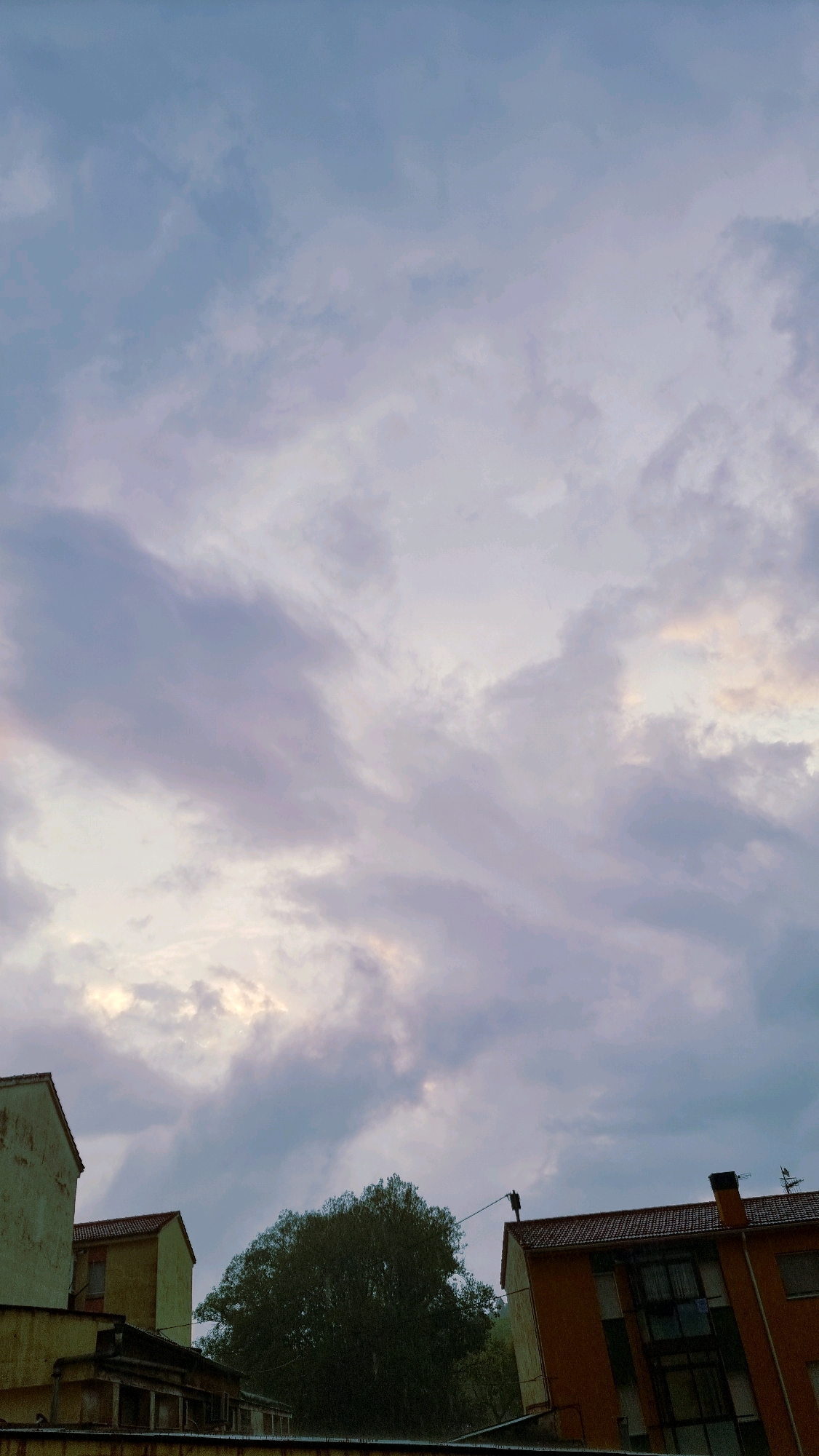 A cloudy sky looms over buildings and a tree in Sant Joan Les Fonts, Spain. The sky displays a gradient of blues and grays with patches of lighter clouds, suggesting either dawn, dusk, or overcast conditions. In the lower portion of the image, several buildings are visible. On the far left, a portion of a light-colored building with a textured facade is present. Adjacent to it, a smaller yellow building with a red tiled roof stands. In the center, a large, dense green tree dominates the midground. To the right of the tree, an orange building with a red tiled roof is prominent, featuring multiple windows. A dark pole or structure extends from the roof of this orange building. Another building, partially visible on the far right, also has a red tiled roof. The overall scene appears to be an urban or semi-urban environment.