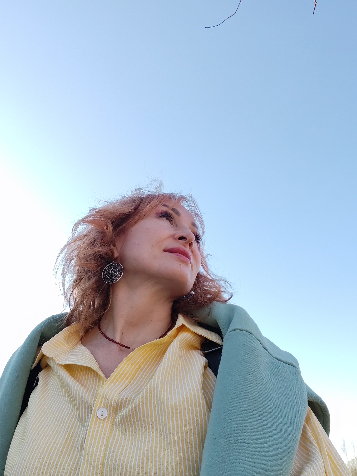 A low-angle shot captures a woman looking upwards against a clear, light blue sky. Her reddish-blonde, wavy hair is tousled by the wind. She wears a yellow and white striped collared shirt, buttoned up, and a light blue sweatshirt or jacket rests on her shoulders. A dark red bead necklace adorns her neck, and she sports a large, circular earring. The location appears to be outdoors. Without additional reference points, I can't confidently ascertain the city of Izhevsk, Russia as the specific location.