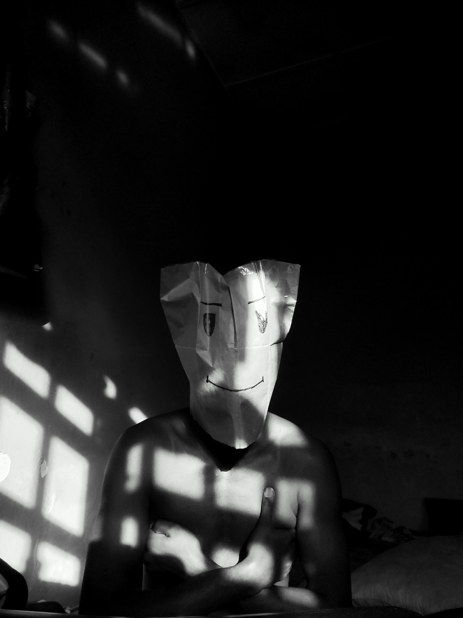 A black and white, low-angle shot depicts a torso of an individual wearing a paper bag over their head with a drawn-on smiley face. The individual is positioned near a window with a grid pattern, creating patterned shadows on their body and the surrounding wall. The individual’s arms are crossed. The image appears to have been taken indoors. There is no verifiable evidence of location in Keffi, Nigeria.